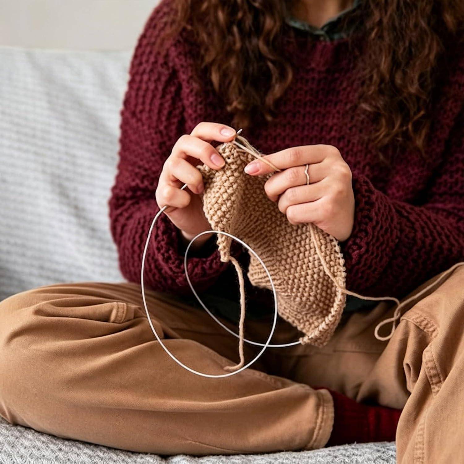 Eine Person strickt ein beiges Stoffstück mit Rundstricknadeln, während sie mit gekreuzten Beinen auf einem Sofa sitzt. Sie trägt einen kastanienbraunen Pullover und braune Hosen.