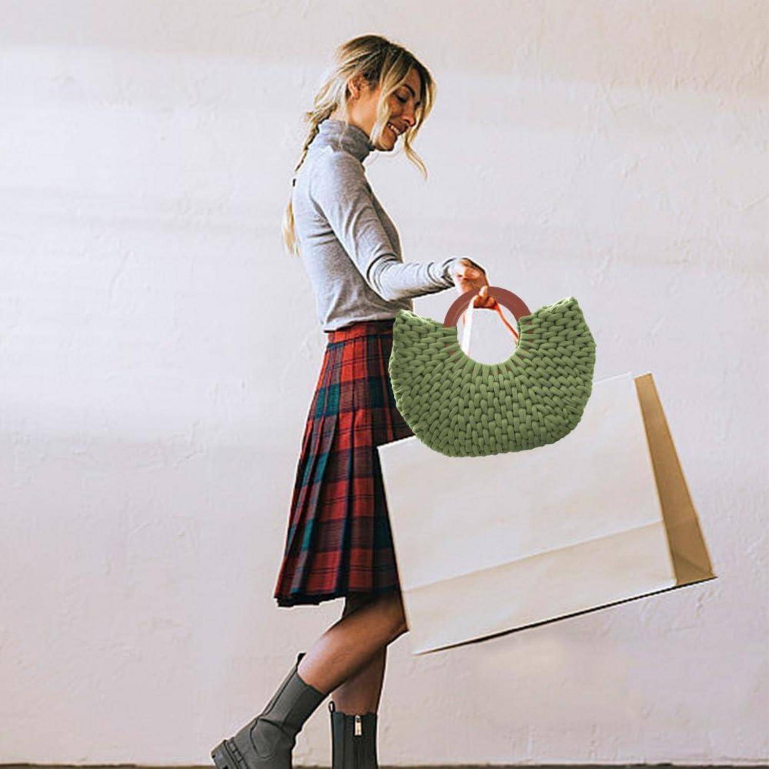 Eine Frau in einem karierten Rock trägt zwei große Einkaufstaschen, darunter eine grüne Webtasche, während sie an einer schlichten Wand entlang geht.