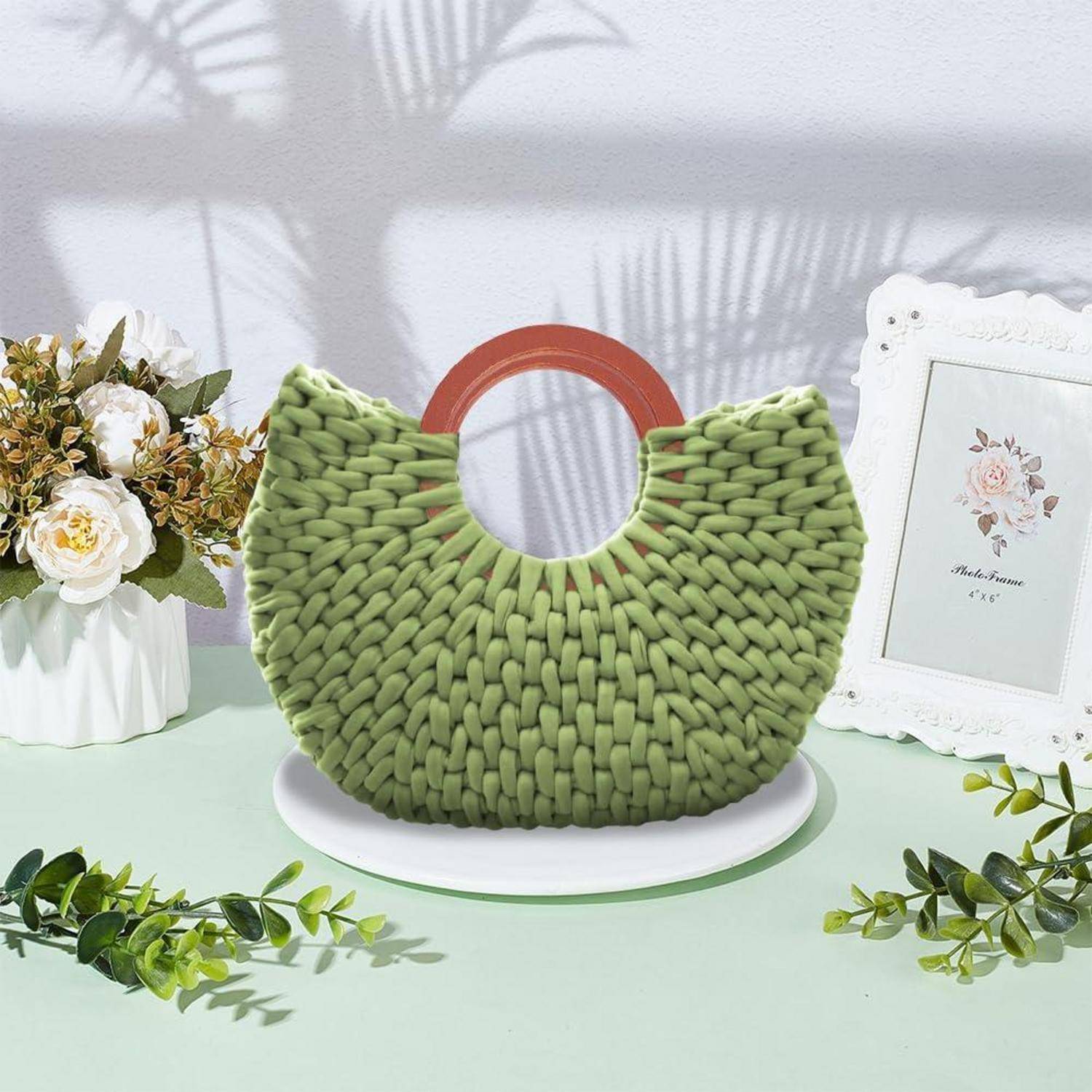 Grüne Woven-Handtasche mit rundem roten Griff, ausgestellt auf einem weißen Ständer neben Blumen und einem gerahmten Druck.