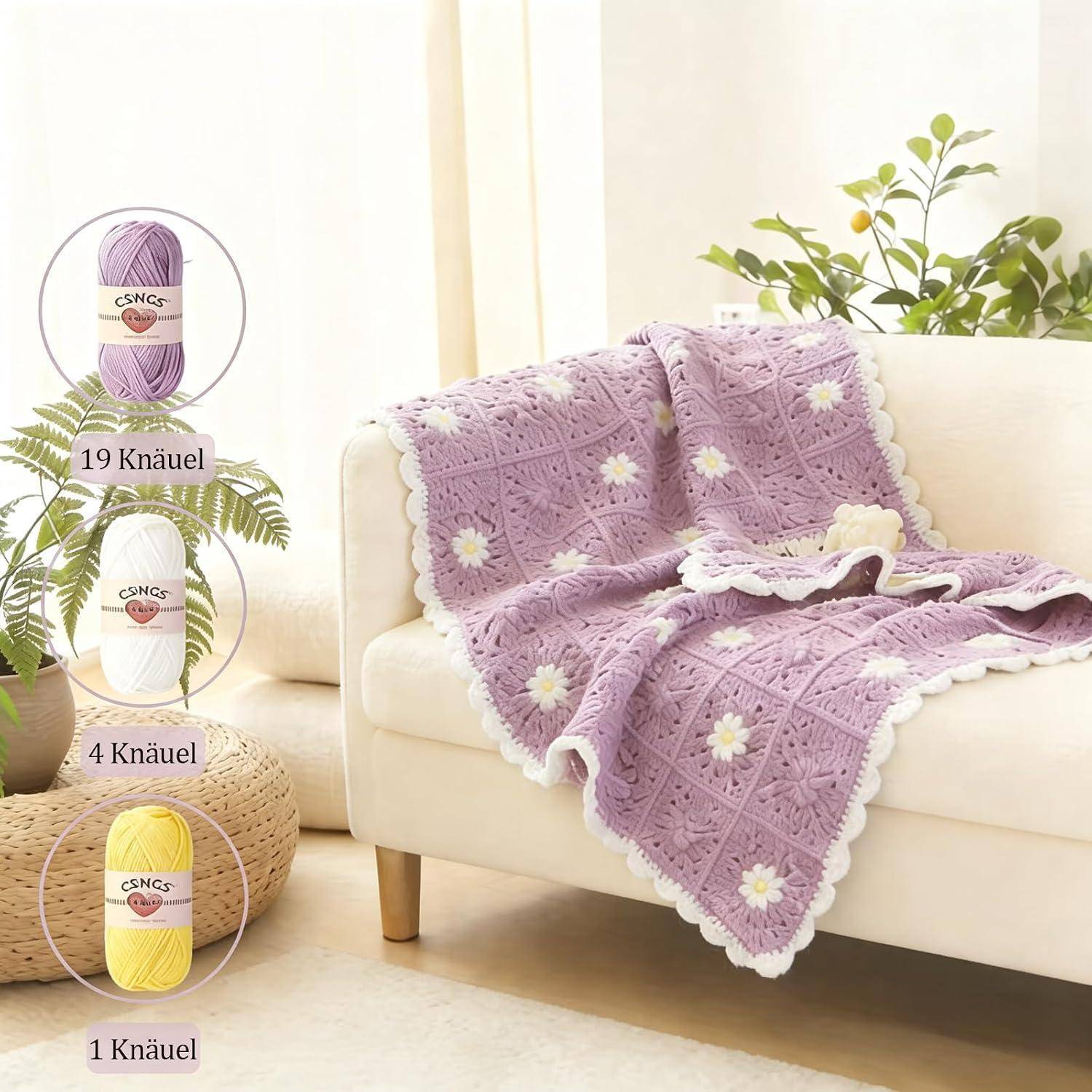 Lavendelgehäkelte Decke mit weißen Gänseblümchen-Quadraten, die über ein cremefarbenes Sofa drapiert sind; eingefügte Wollknäuel mit den Etiketten „19 Knäuel