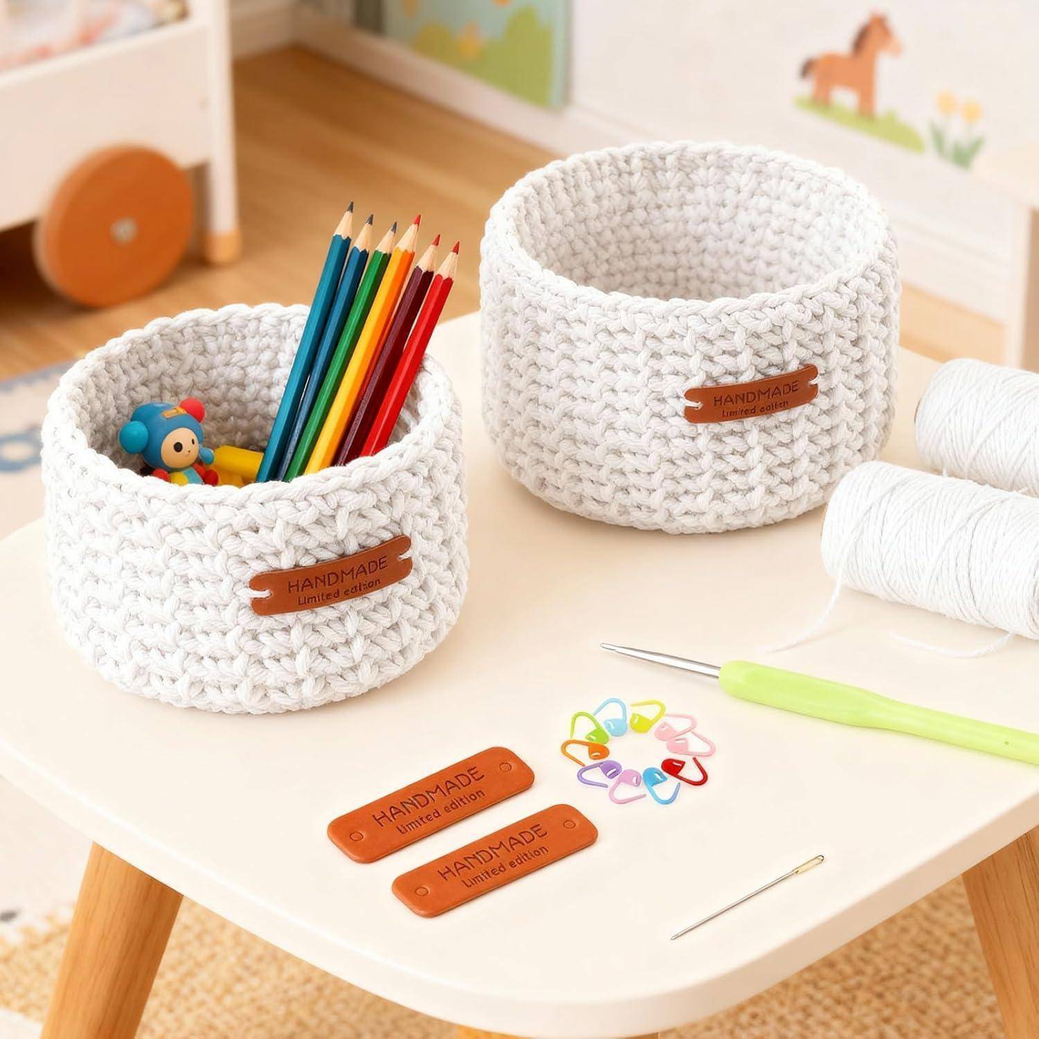Zwei weiße gehäkelte Aufbewahrungskörbe auf einem Tisch, einer mit Buntstiften und Spielzeug gefüllt, mit handgefertigten Lederetikett und Handwerkzeugen in der Nähe.
