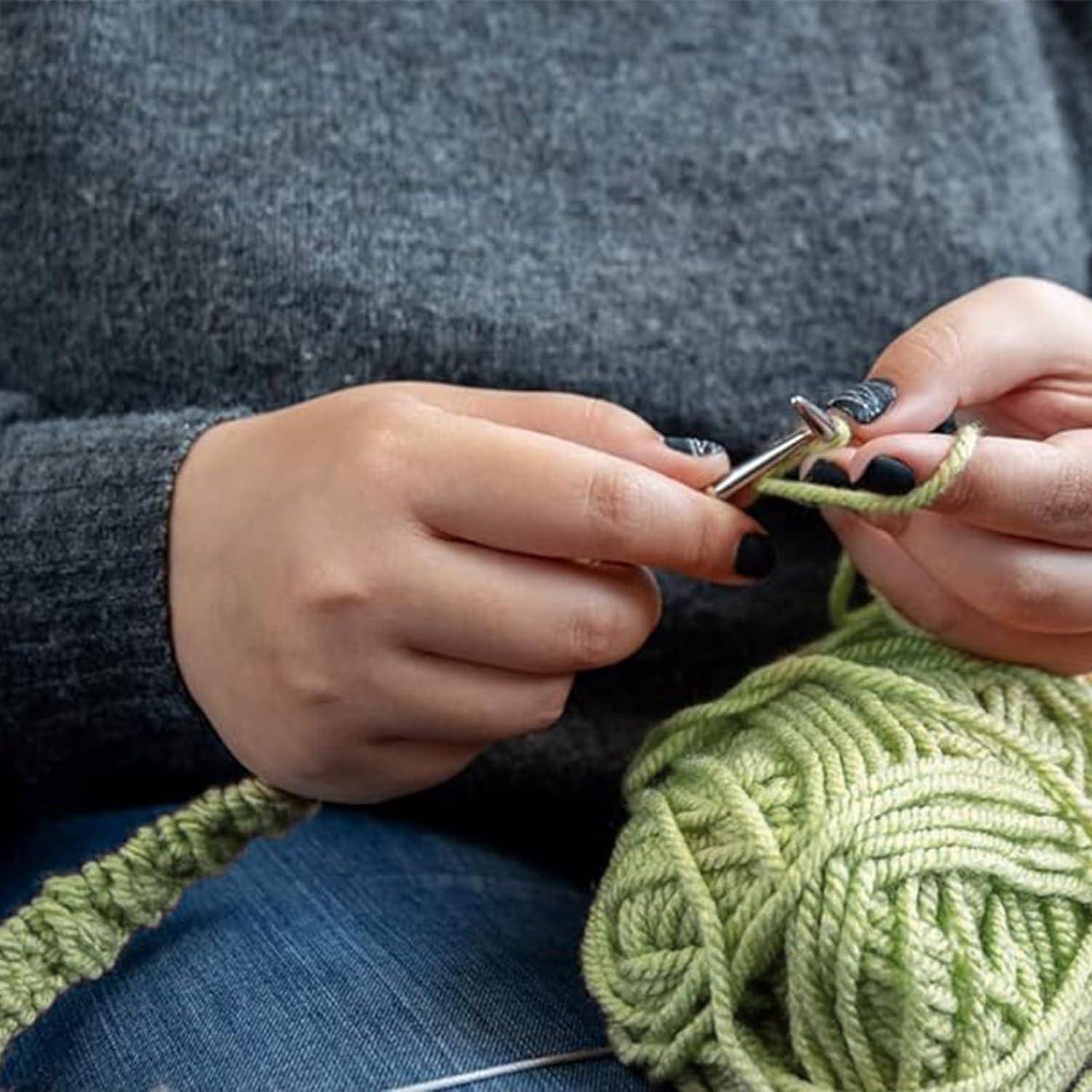 Nahaufnahme von Händen, die mit grünem Garn und Metallnadeln stricken, mit einem Wollknäuel auf dem Schoß.