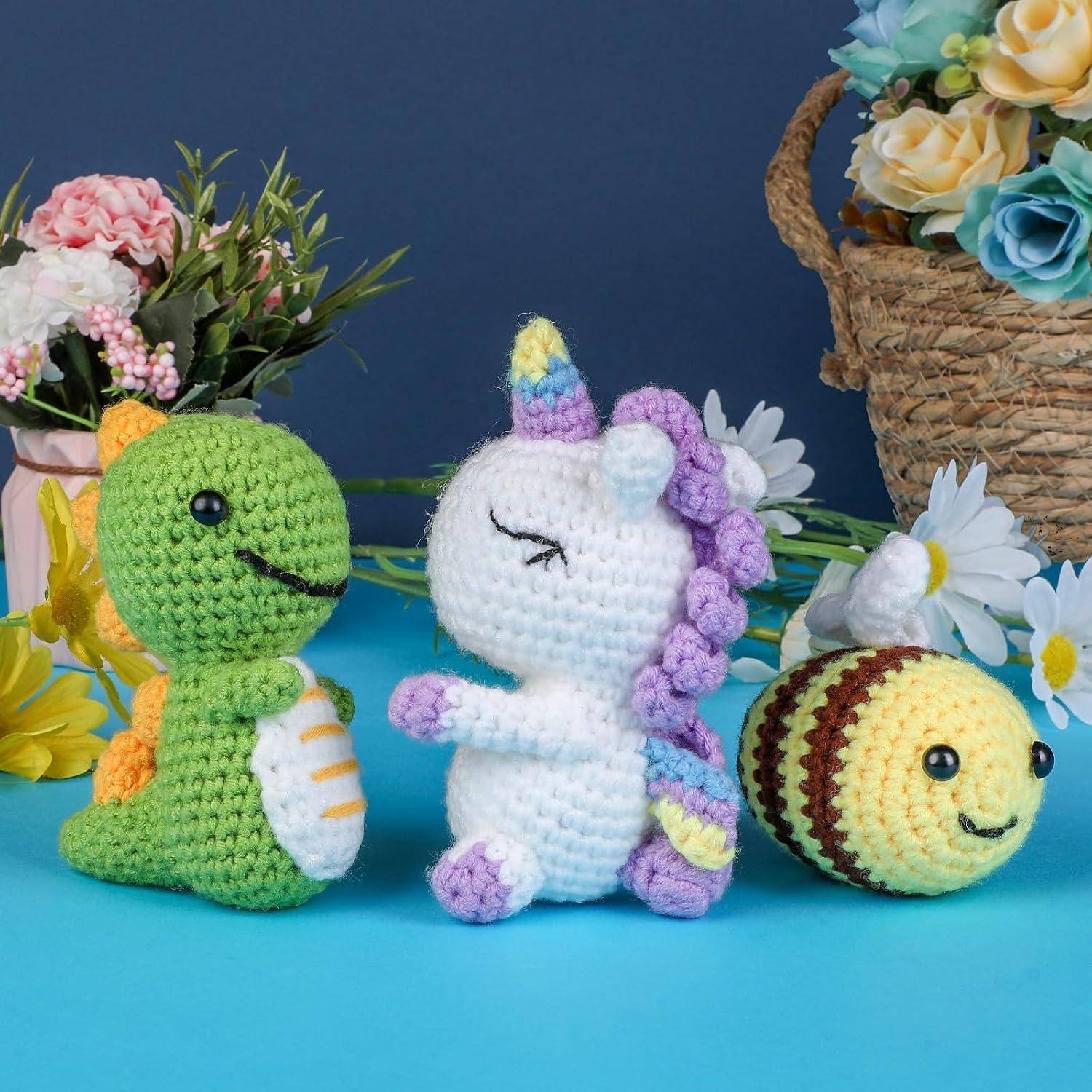 Drei gehäkelte Amigurumi-Spielzeuge auf einer blauen Oberfläche: ein grüner Dinosaurier, ein weißes Einhorn mit Pastellmähne und eine Biene, umgeben von Blumen.