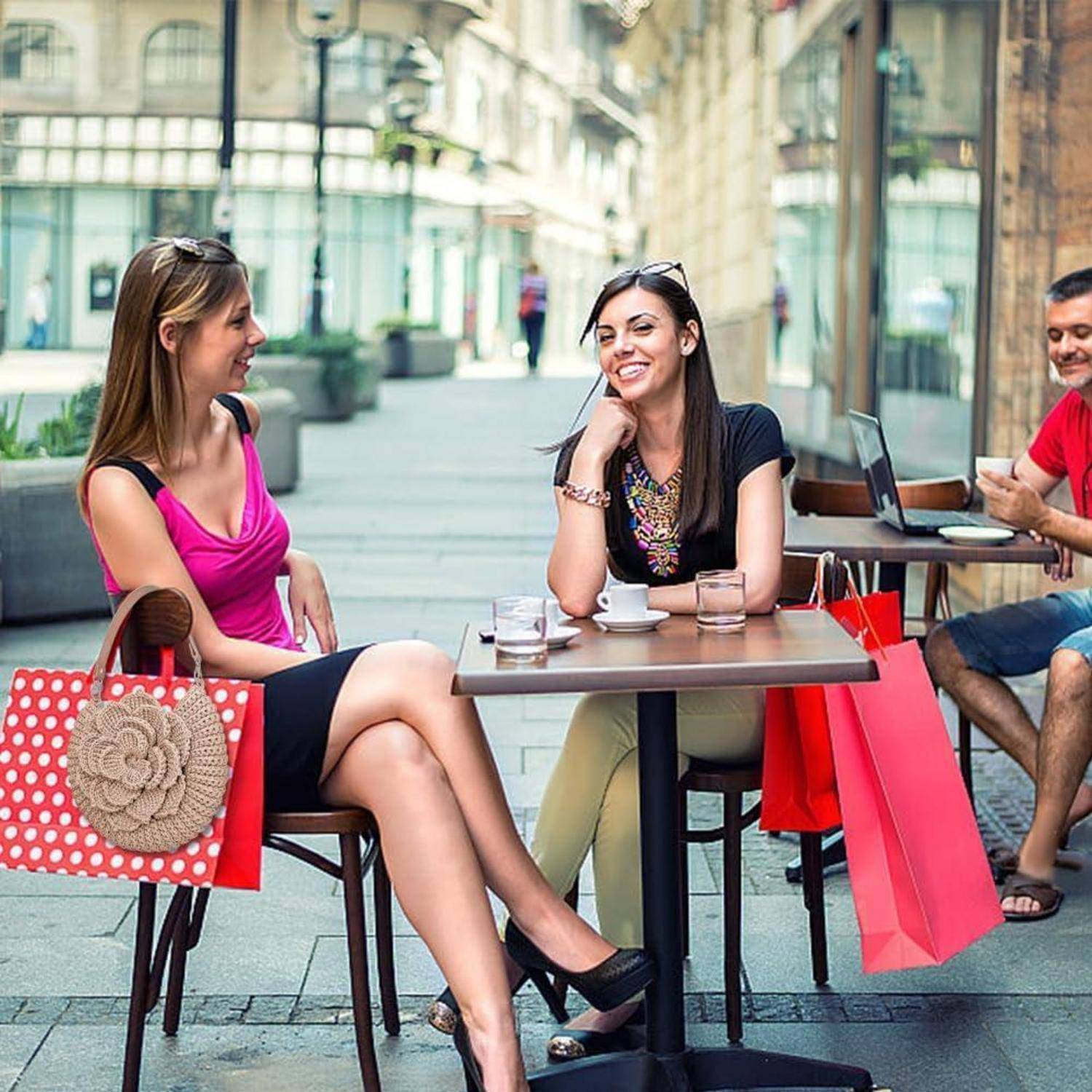 Zwei Frauen unterhalten sich an einem Außencafé mit Einkaufstaschen auf ihren Stühlen, während ein Mann in der Nähe sitzt und einen Laptop und ein Telefon benutzt.