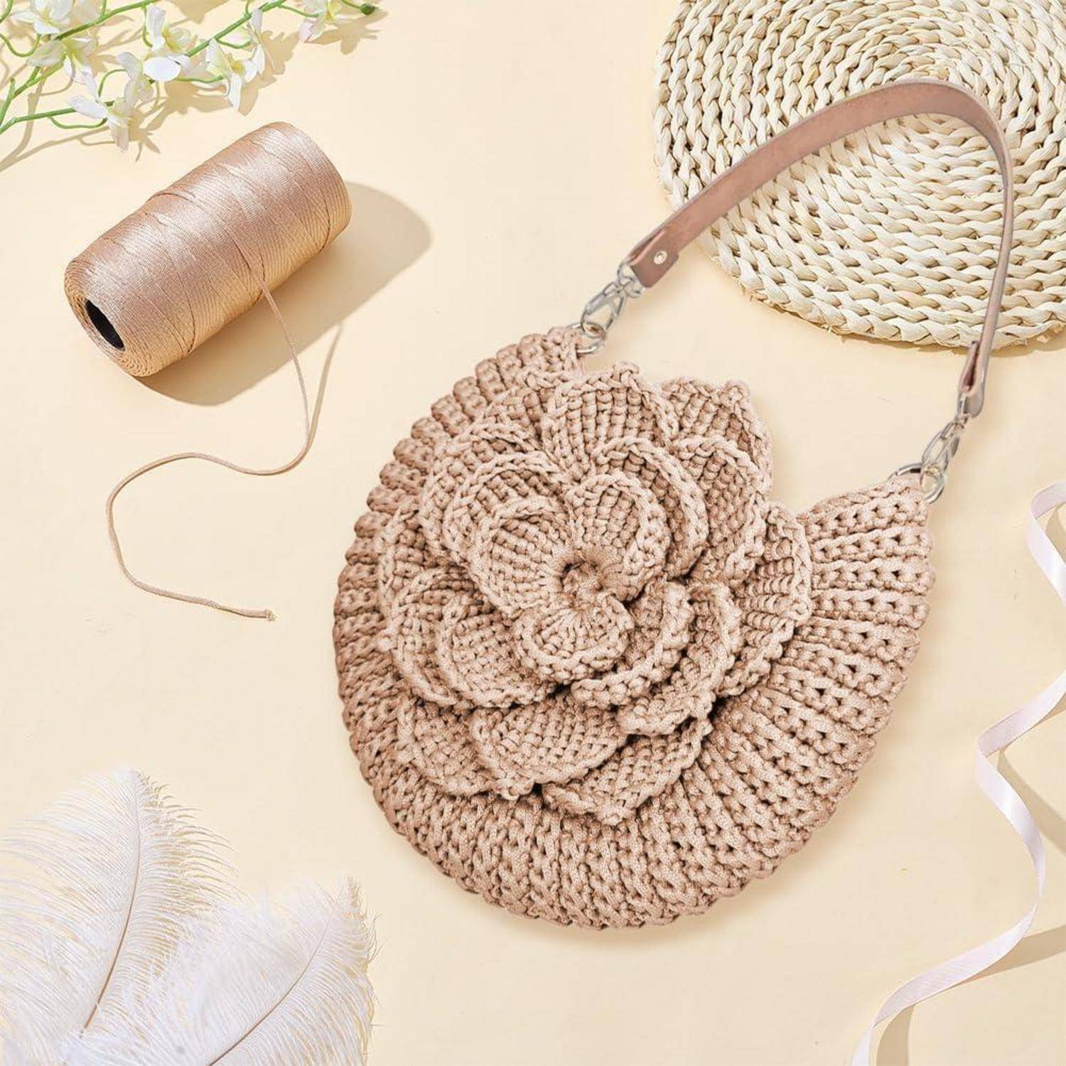 Gehäkelte runde beige Handtasche mit großer Blumenverzierung, gezeigt mit einer Garnrolle und gewebtem Hut auf hellem Hintergrund.