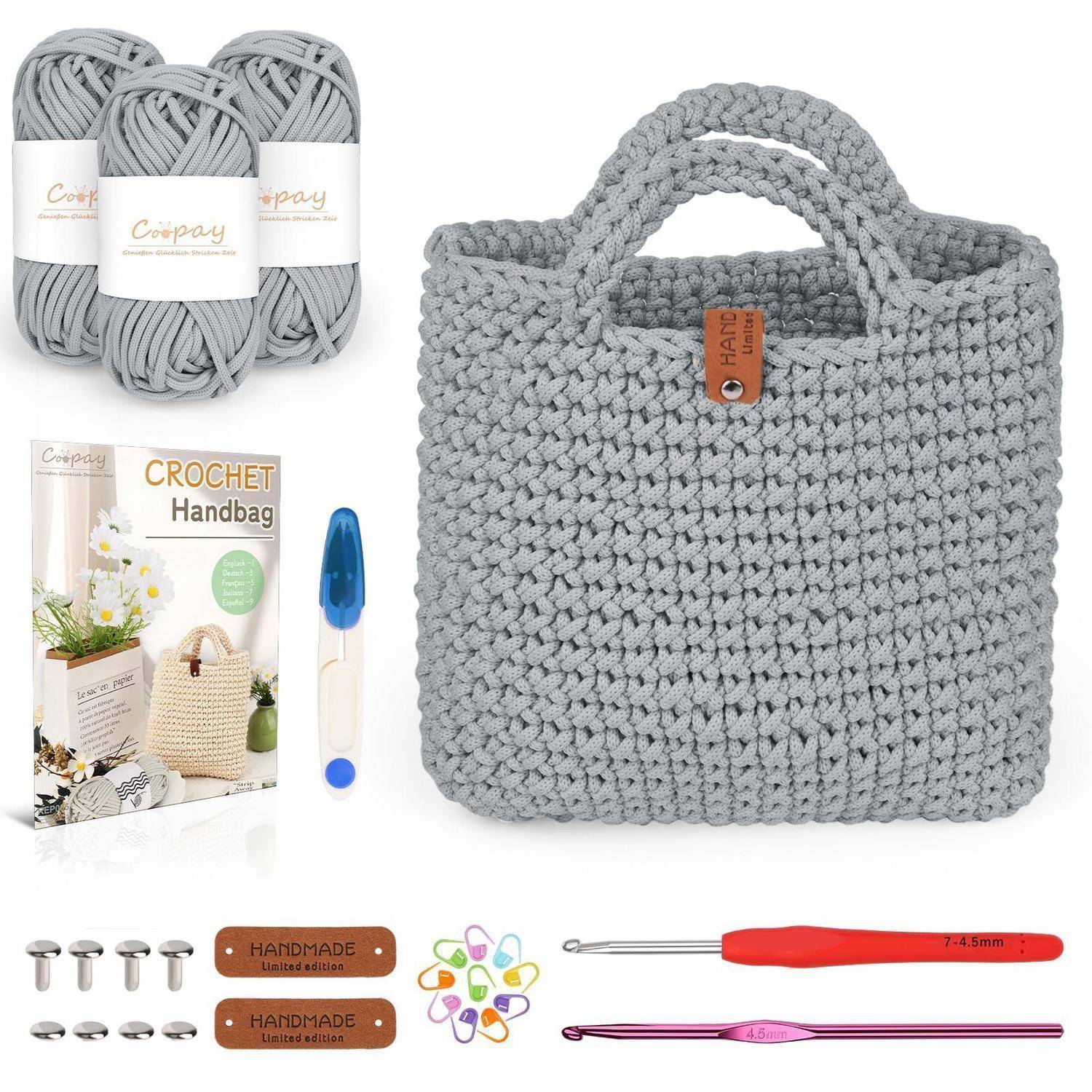 Grau gehäkelte Handtasche Set mit Garn, Musterbroschüre, Häkelnadel, Zubehör und fertige Tasche auf weißem Hintergrund.