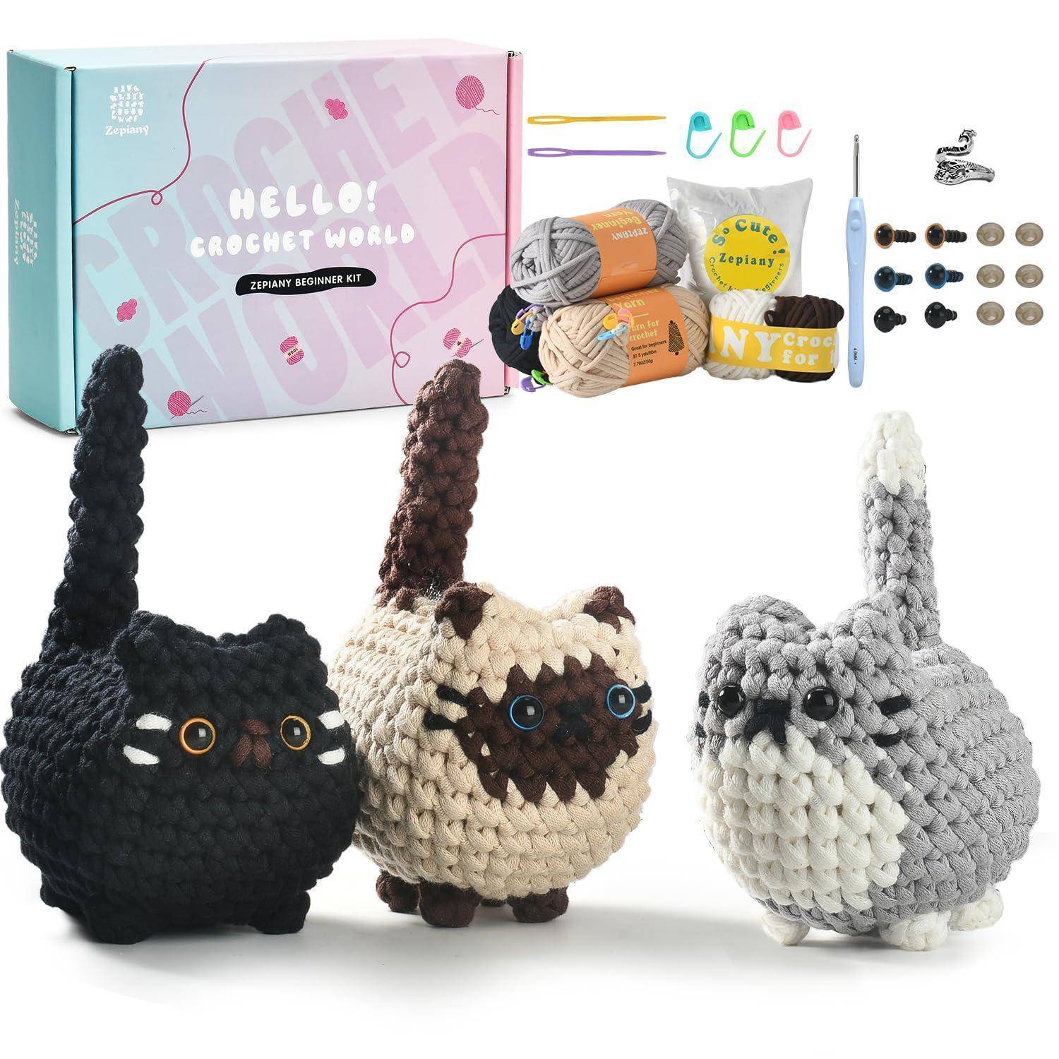 Häkelset für Anfänger mit Garn, Häkelhaken und Zubehör, gezeigt mit drei fertigen Katzenfiguren in Schwarz, Braun-Creme und Grau-Weiß.