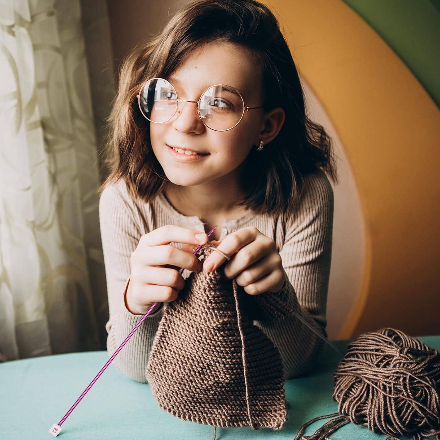 Junges Mädchen mit runder Brille, das an einem Tisch eine braune Tasche mit rosa Nadeln strickt und dabei Garn neben einem Wollknäuel hält.