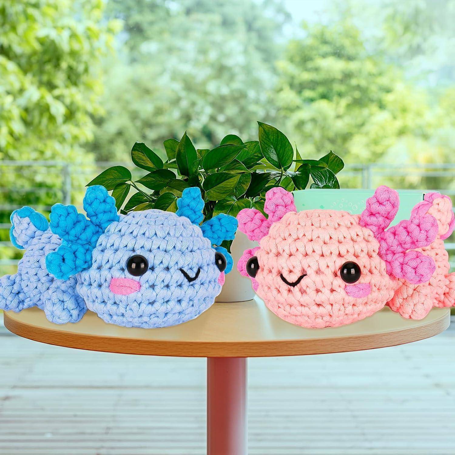 Zwei gehäkelte Axolotl-Kuscheltiere, eines blau und eines rosa, sitzen auf einem kleinen runden Tisch mit einer Topfpflanze hinter ihnen.