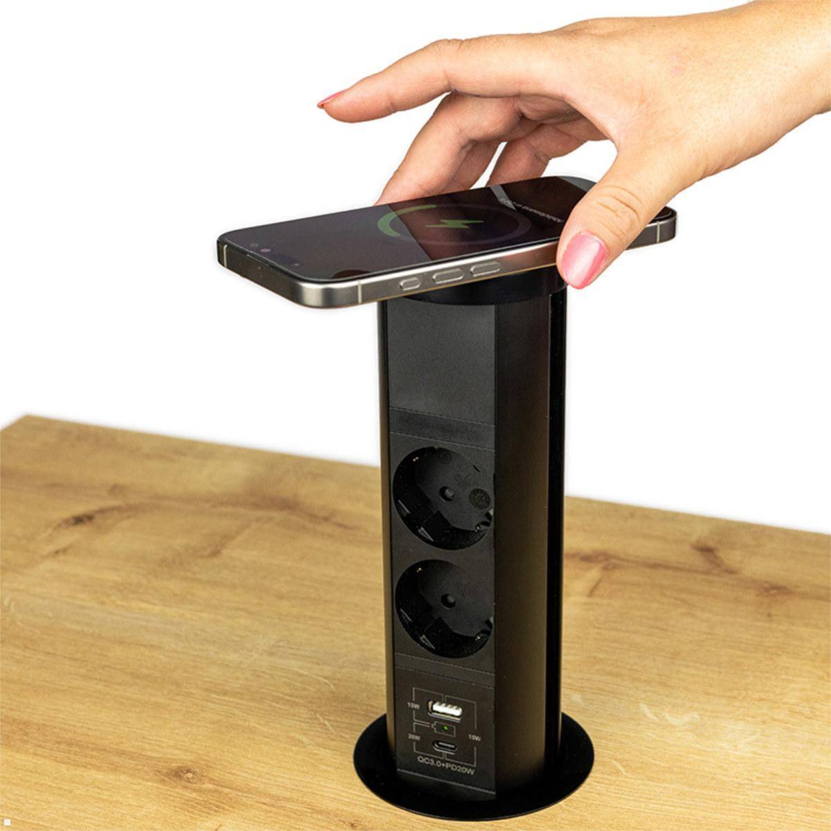 Hand, die ein Smartphone auf einen vertikalen Pop-up-Schreibtisch-Stromturm mit Steckdosen und USB-Anschlüssen auf einem Holztisch platziert.