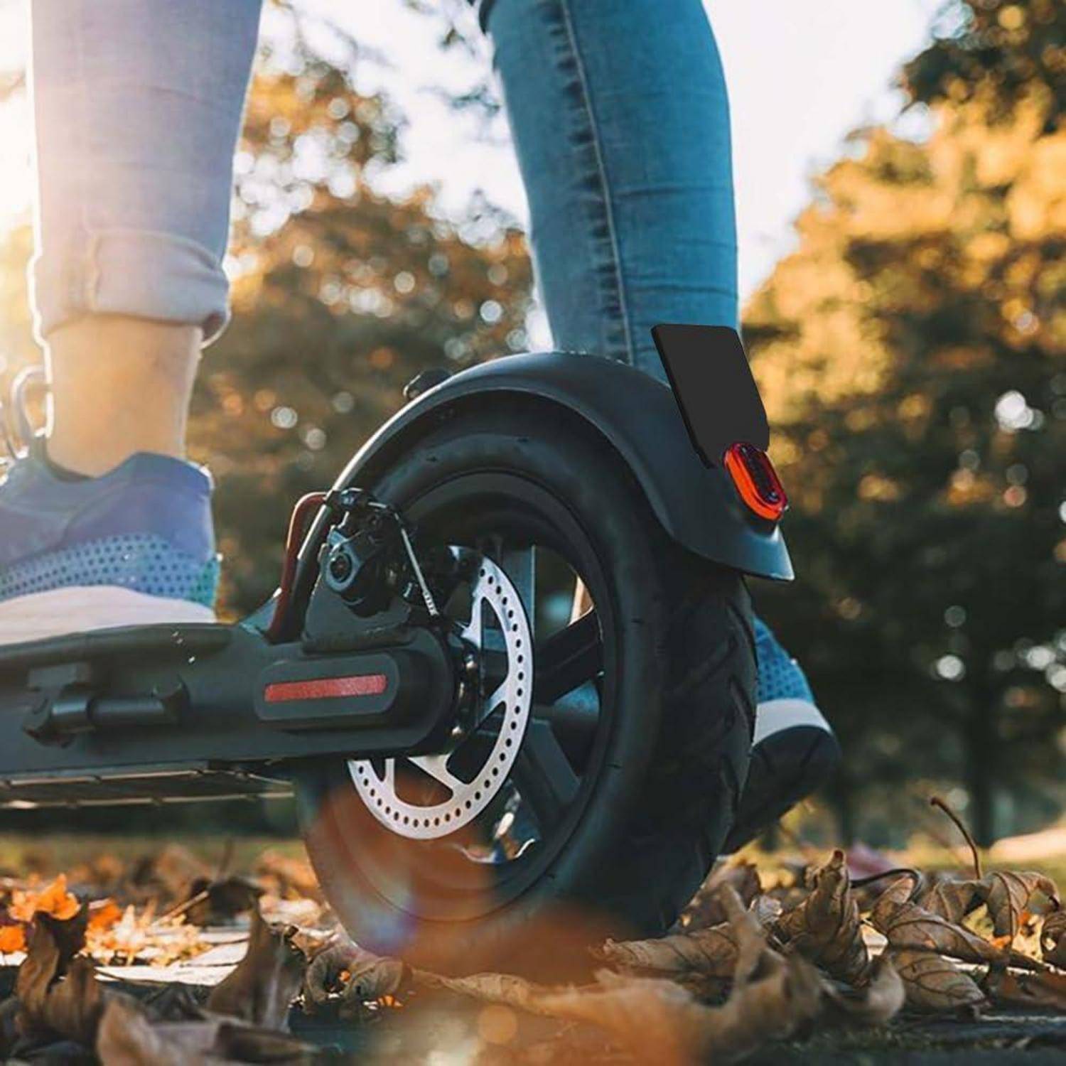 Nahaufnahme eines elektrischen Rollerrades, das über gefallene Blätter rollt, mit sichtbaren Beinen des Fahrers draußen bei Sonnenuntergang.