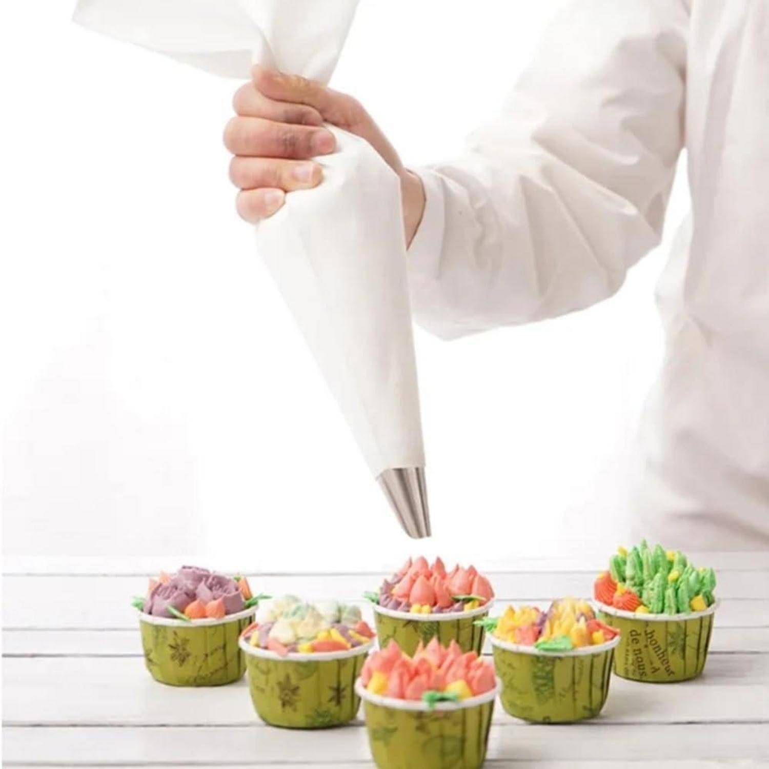 Eine Person spritz Zuckerguss aus einem Spritzbeutel auf mehrere bunte Cupcakes in grünen Papierförmchen auf einer weißen Oberfläche.