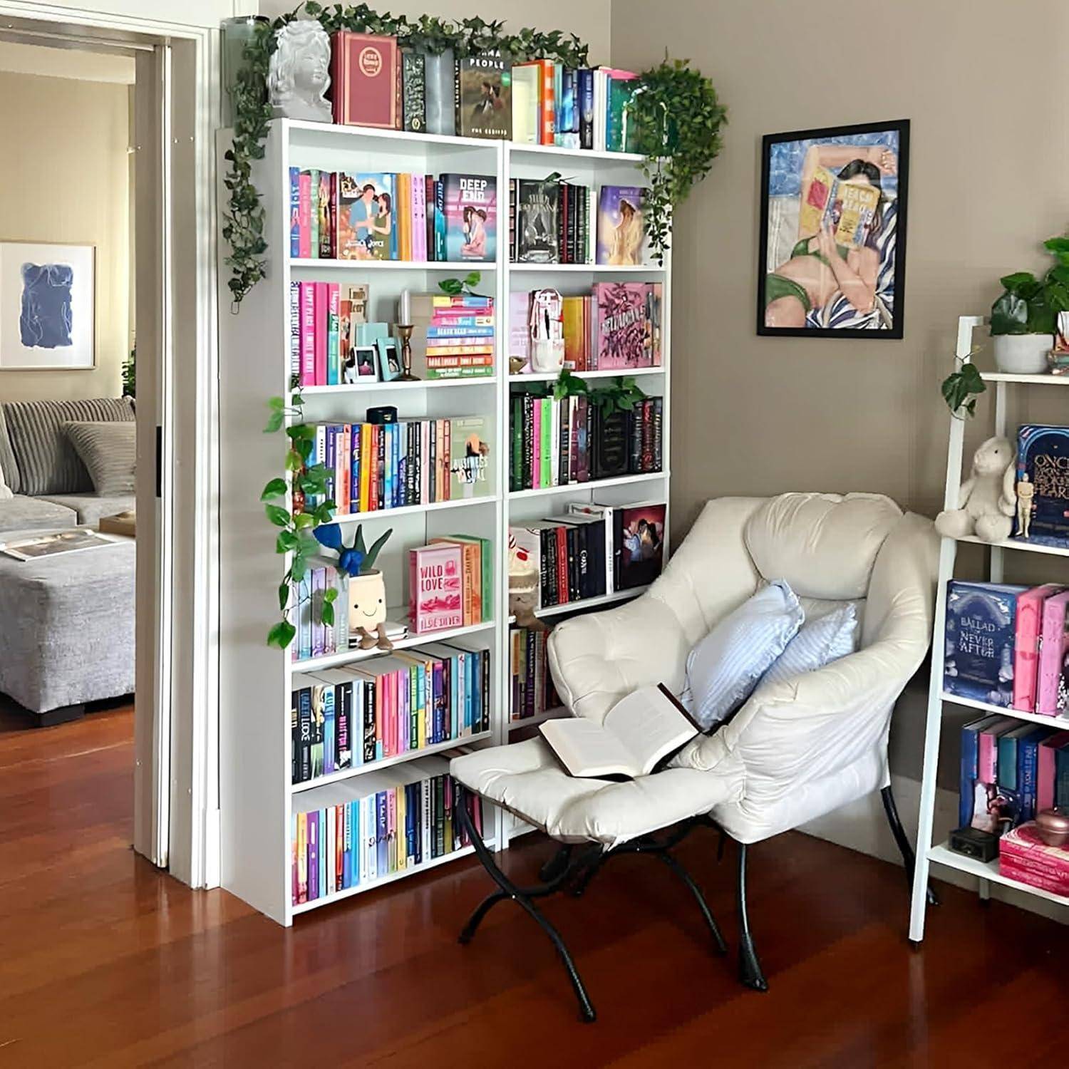 Gemütliche Leseecke mit einem weißen Bücherregal voller bunter Bücher, einem cremefarbenen Loungestuhl mit einem aufgeschlagenem Buch und gerahmten Wandbildern.