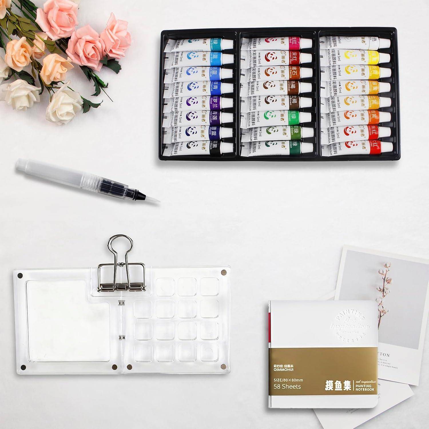 Aquarellmalkasten mit Pinsel, künstliche Blumen, leeres Binderblatt und kleines Notizbuch auf einer weißen Tischplatte.