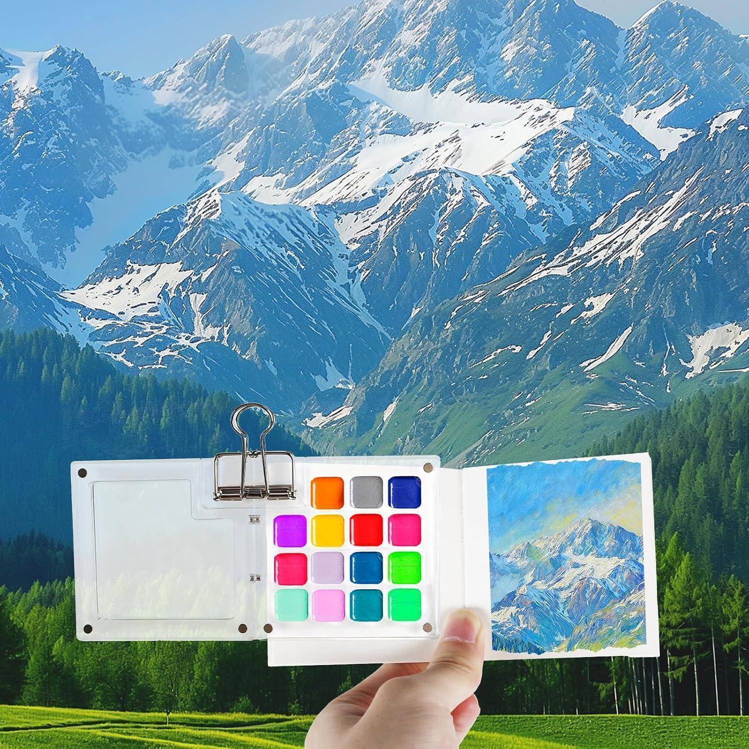 Hand, die eine kleine Aquarellpalette und ein Malblock vor schneebedeckten Bergen und Wald hält, zeigt eine Miniaturlandschaftsmalerei in Bearbeitung.