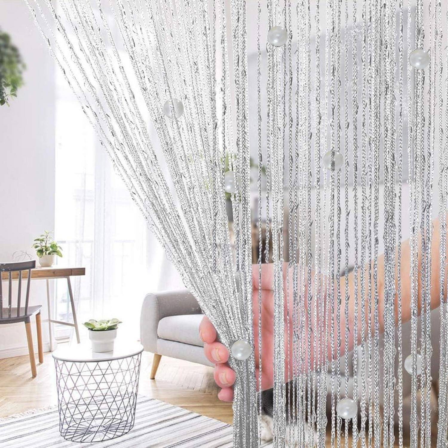 Hand, die ein silbernes Perlenvorhang-Panel in einem hellen Wohnzimmer hält, zeigt einen dekorativen Raumteiler oder Fensteraufhänger.