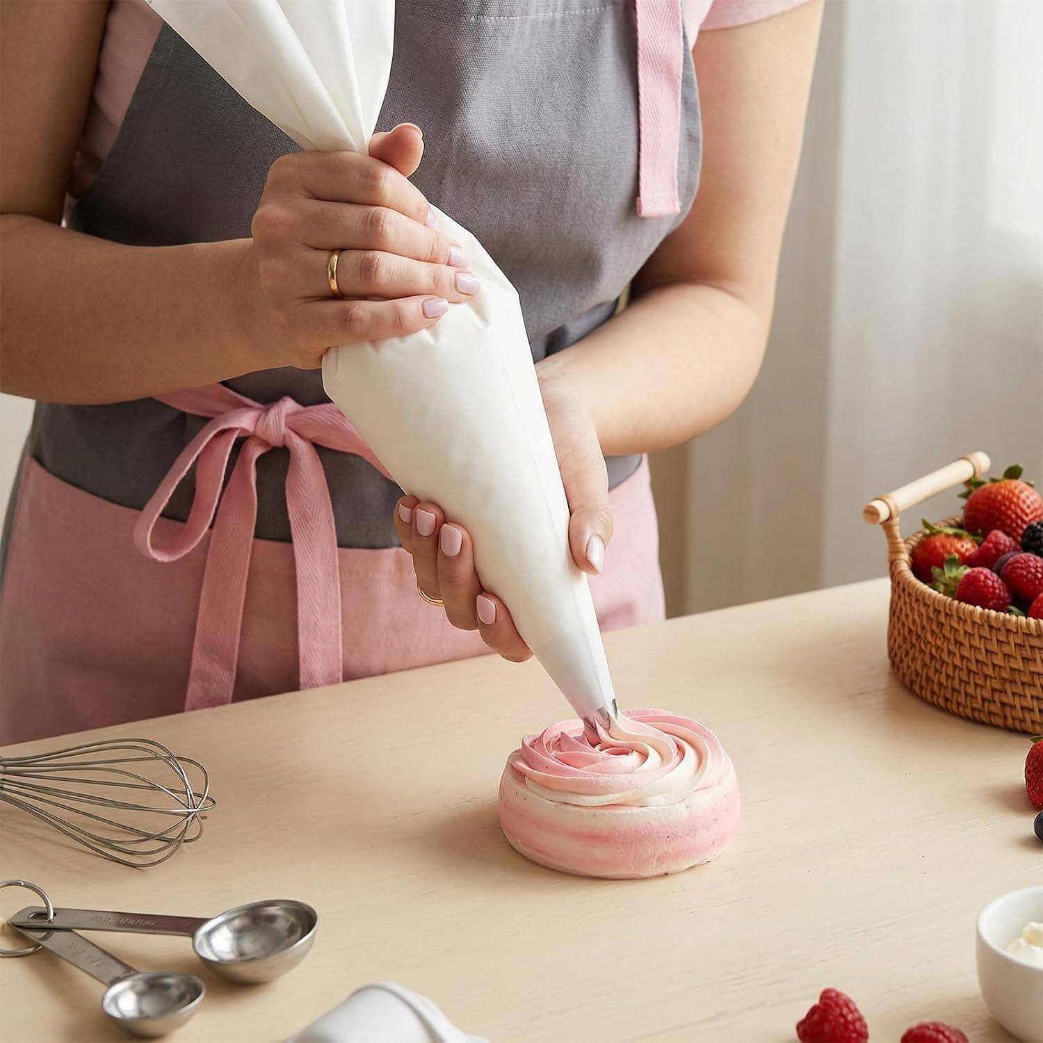 Person, die rosa Zuckerguss mit einem Spritzbeutel auf einen kleinen Kuchen oder ein Dessert auf einer Küchentheke aufträgt, mit Backwerkzeugen und Erdbeeren in der Nähe.