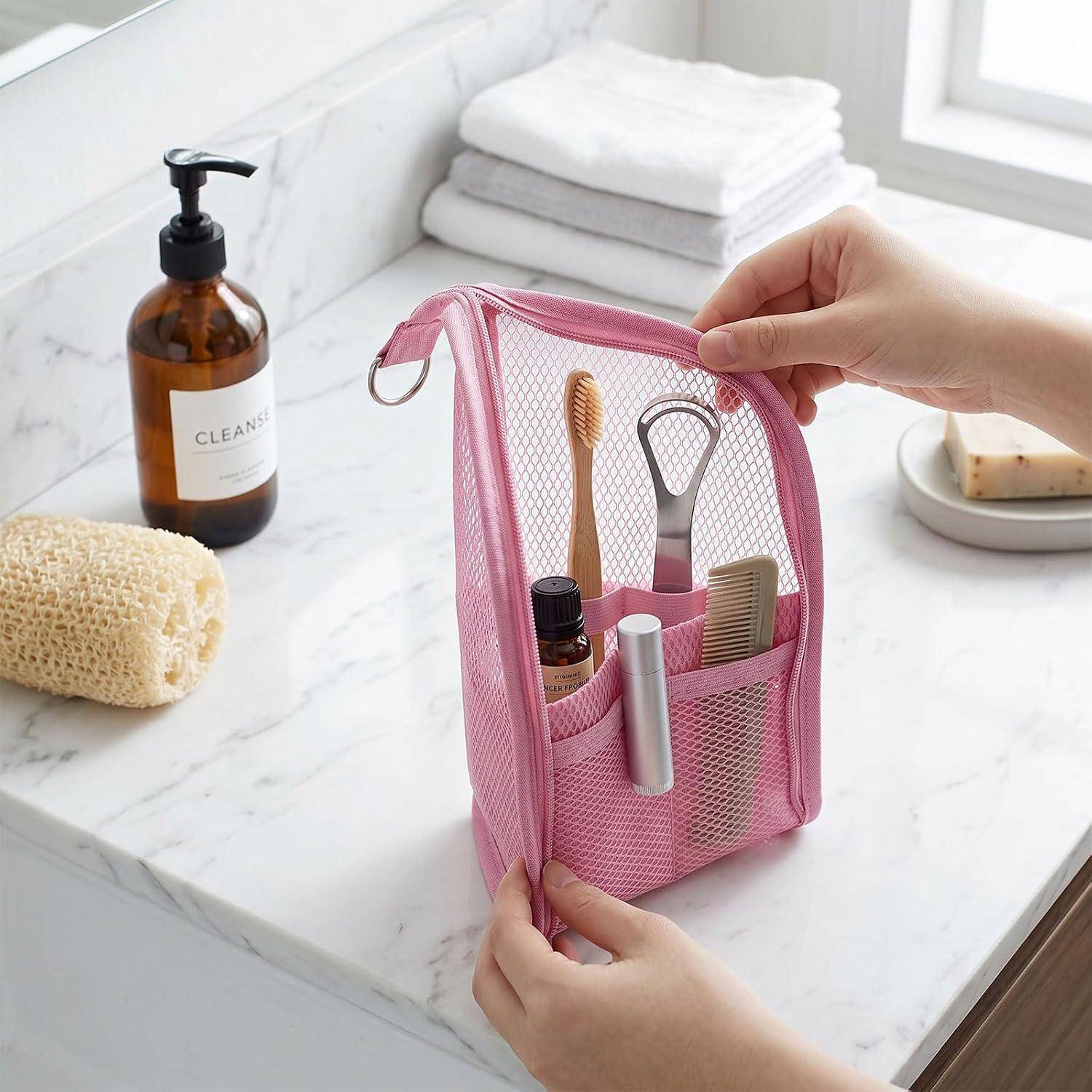 Hände halten einen rosa Mesh-Kosmetiktaschen auf einer Badezimmer-Theke, zeigen Zahnbürste, Rasierer, Kamm und kleine Flaschen neben gefalteten Handtüchern und Seifenspender.