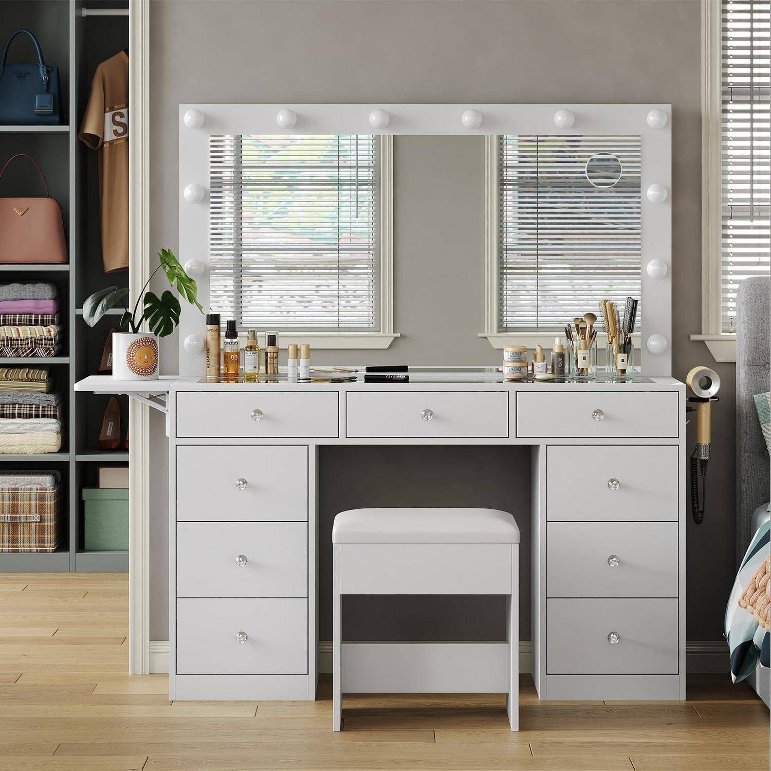Grauer Frisiertisch mit beleuchtetem Spiegel, Make-up und Pinseln oben, und ein weißer Hocker darunter zentriert in einem ordentlichen Schlafzimmer oder Ankleidezimmer.