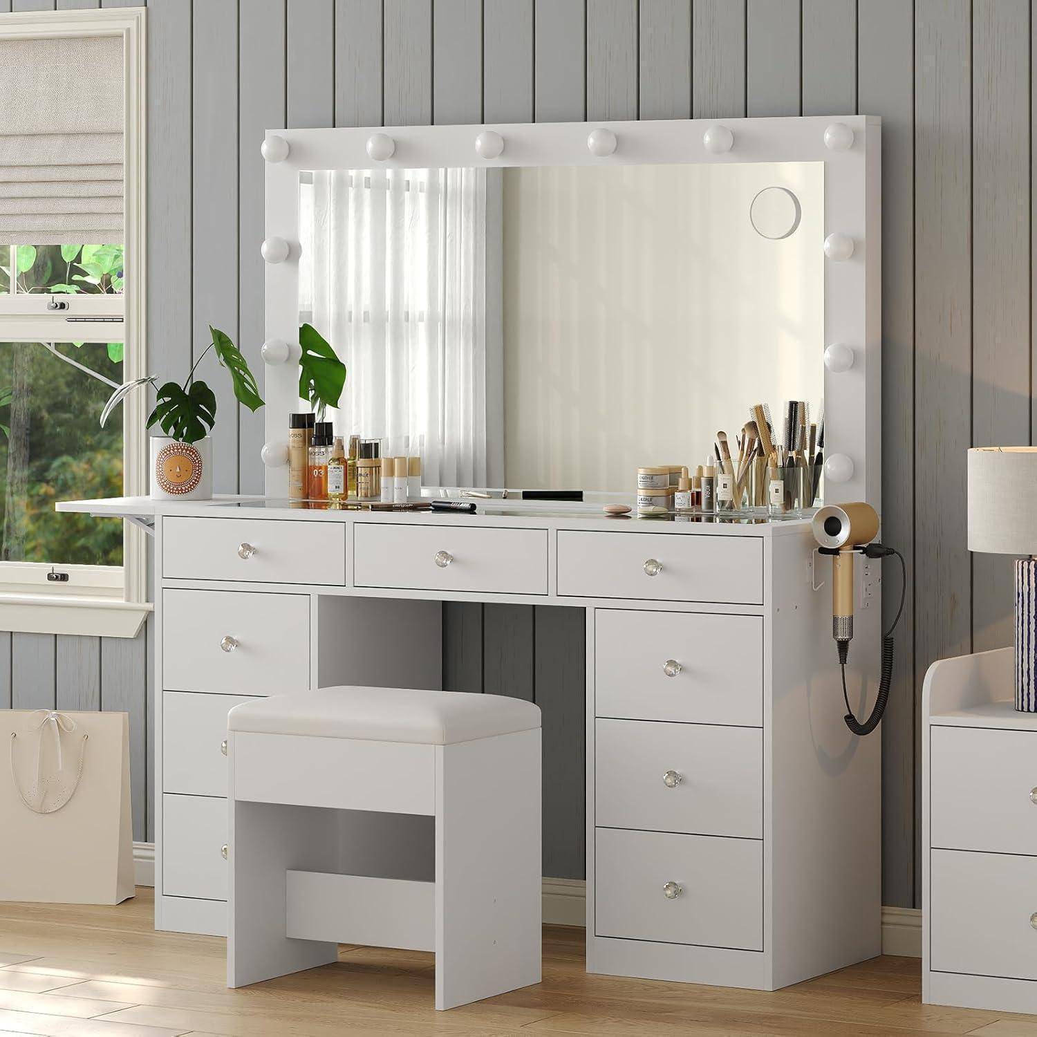 Weißer Schminktisch mit beleuchtetem Spiegel, Schubladen, Hocker und Haarstyling-Geräten in einem hellen Schlafzimmer neben einem Fenster und einer Lampe.