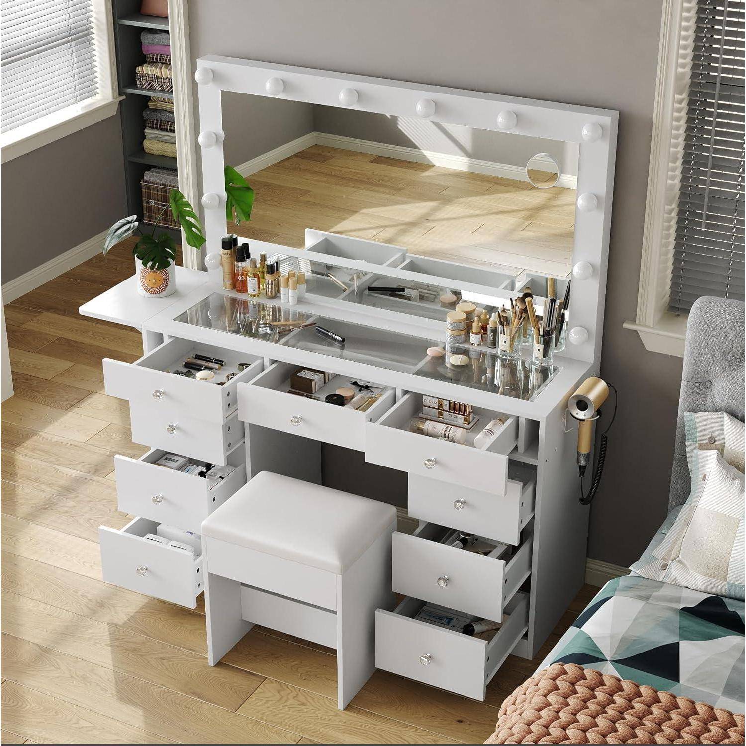Weiße Schminktischkommode mit beleuchtetem Spiegel, offene Schubladen voller Kosmetika und ein Hocker in einem hellen Zimmer neben einem Fenster.