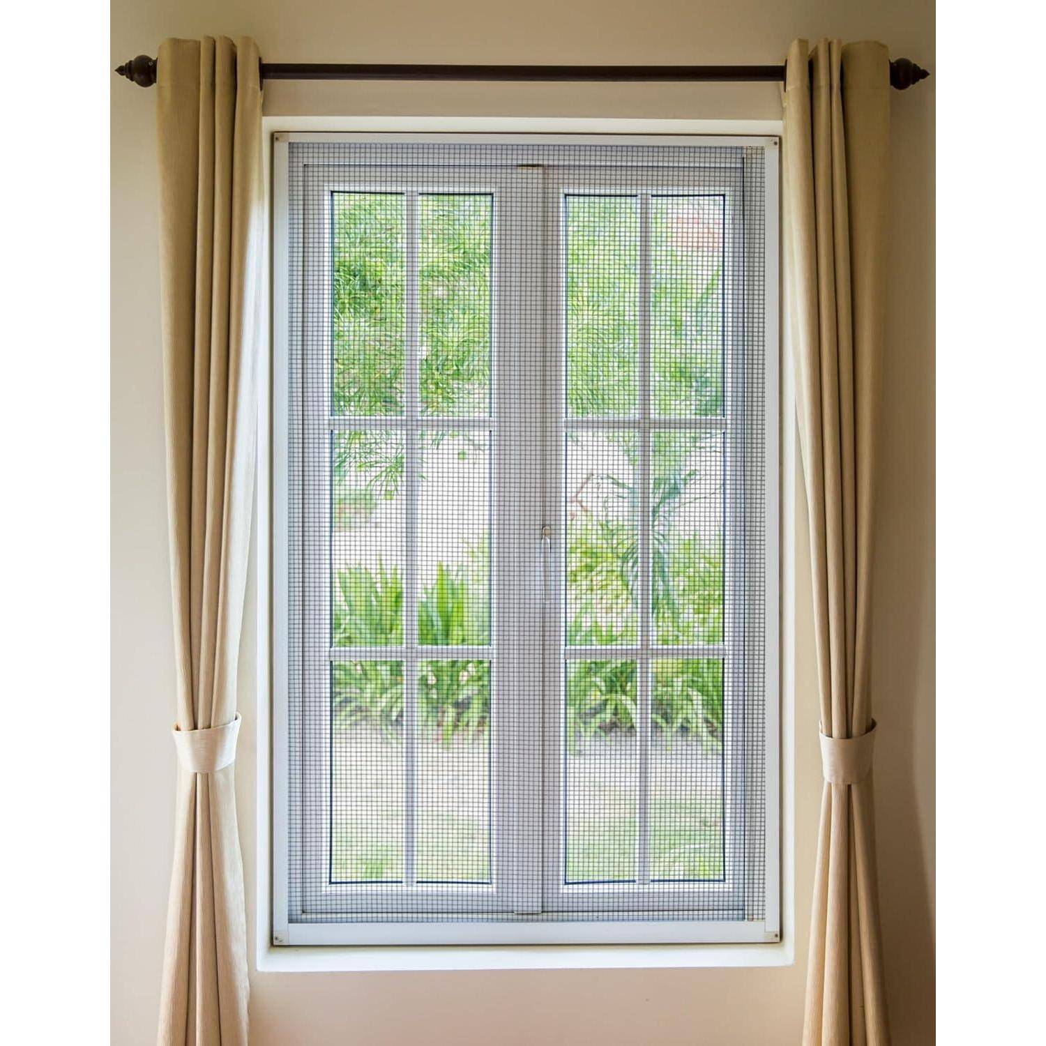 Ein weißes Doppelfenster mit Fliegengitter und beigen Vorhängen, die zur Seite gezogen sind und grünes Grün draußen zeigen.
