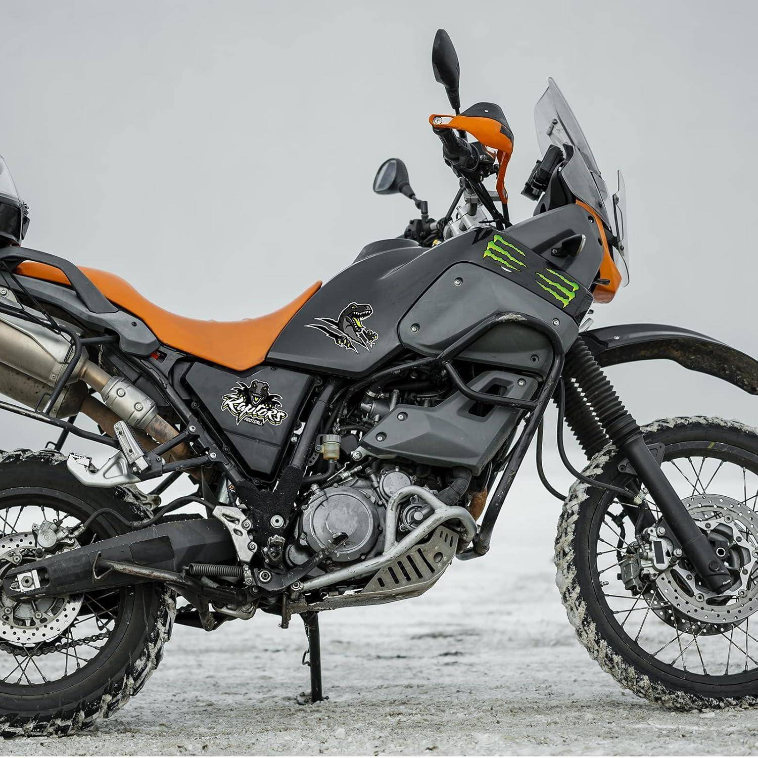 Orange-schwarzes Adventure-Motorrad auf schneebedecktem Boden geparkt, in Seitenansicht mit hohem Windschirm und Stollenreifen dargestellt.