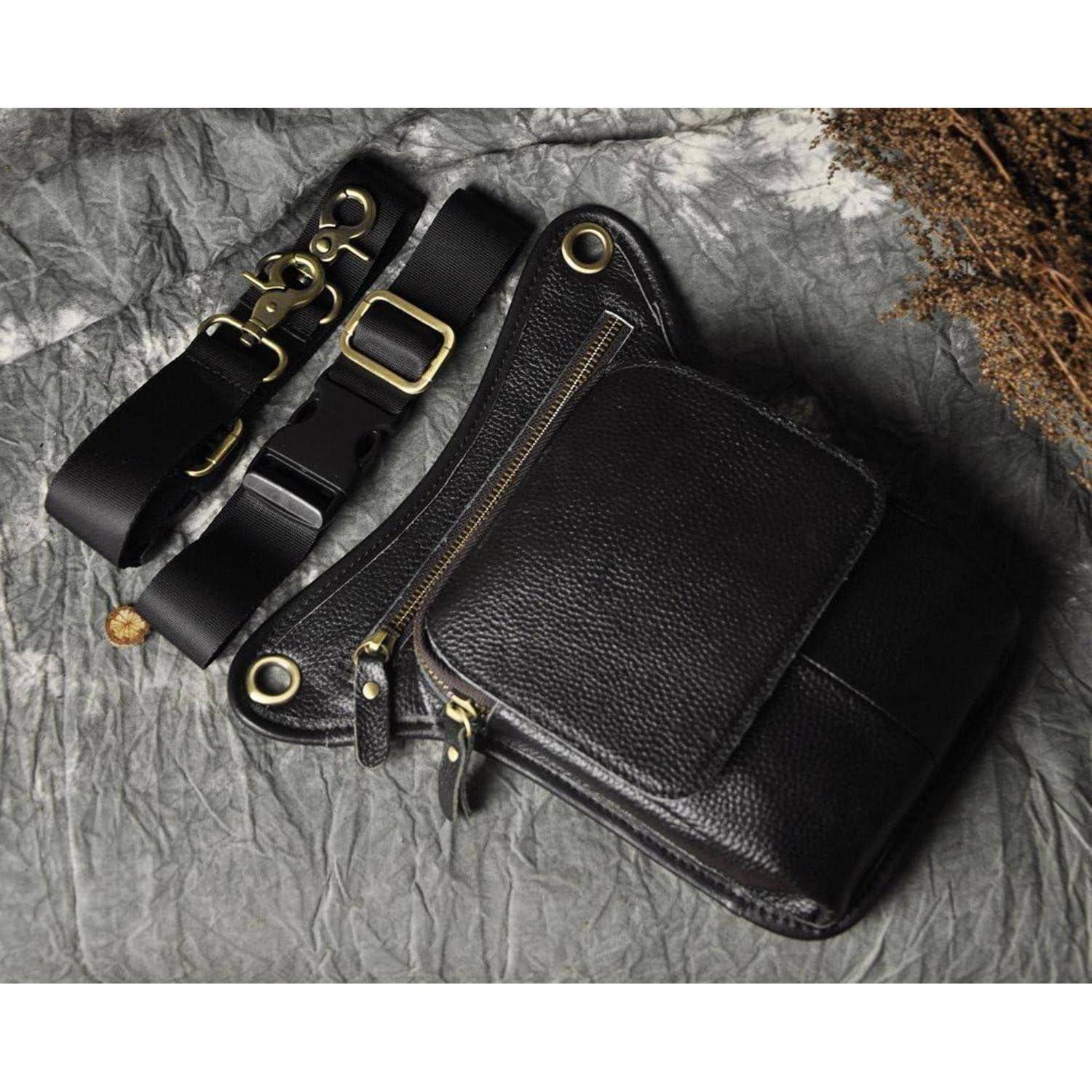 Schwarze Ledertasche zum Umhängen mit goldfarbenem Beschlag, verstellbarem Riemen und Vordertasche mit Klappe, auf grauem strukturiertem Stoffhintergrund gezeigt.