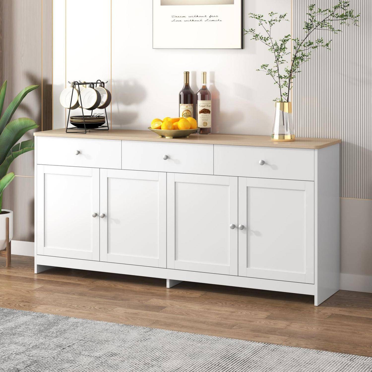 Weißer Sideboard-Schrank mit drei Schubladen und vier Türen, oben dekoriert mit einer Obstschale, Weinflaschen, einer Lampe und einer Vase in einem hellen modernen Raum.