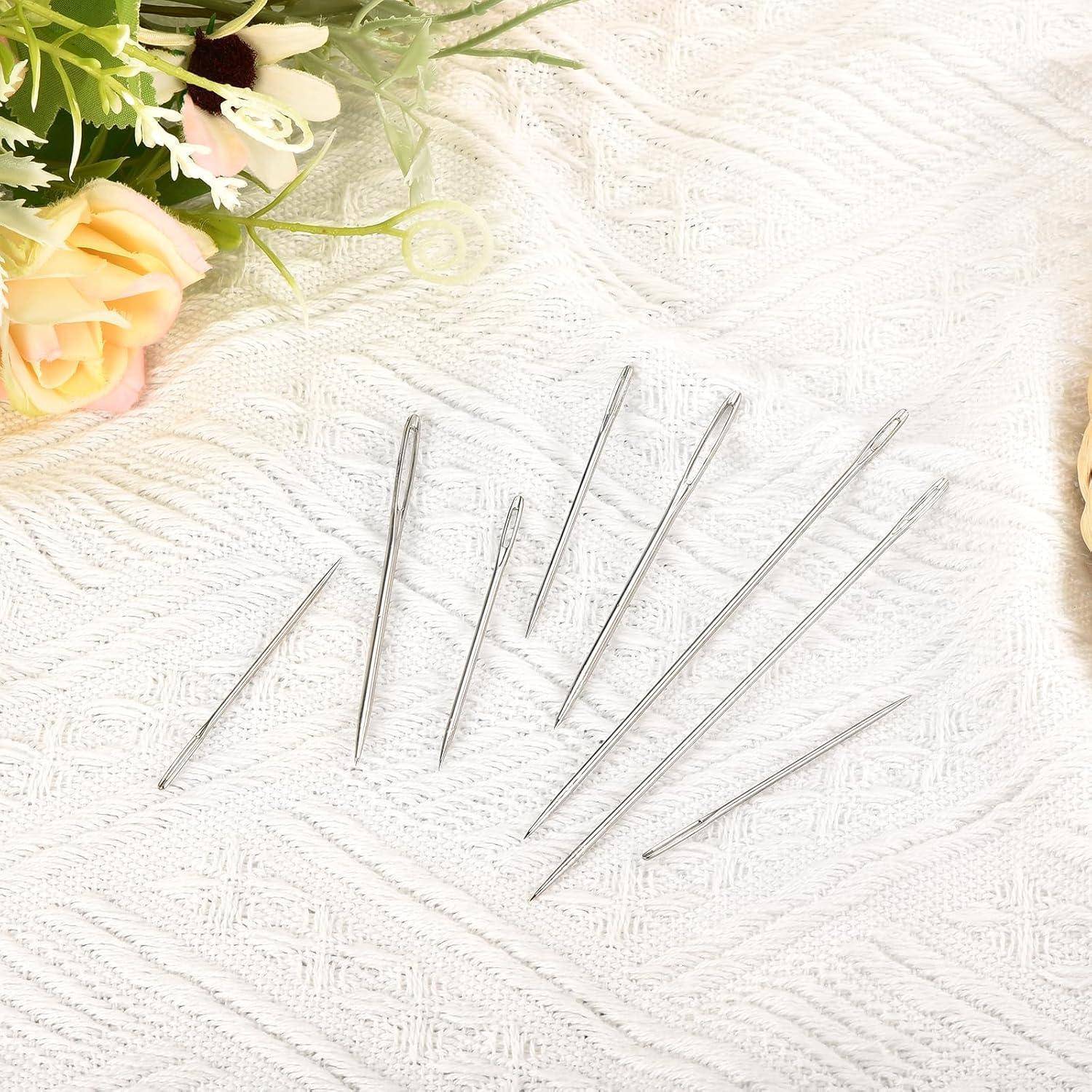 Assortierte silberne Nähnadeln auf einem weißen strukturierten Stoff ausgelegt, mit Blumen in der Ecke.