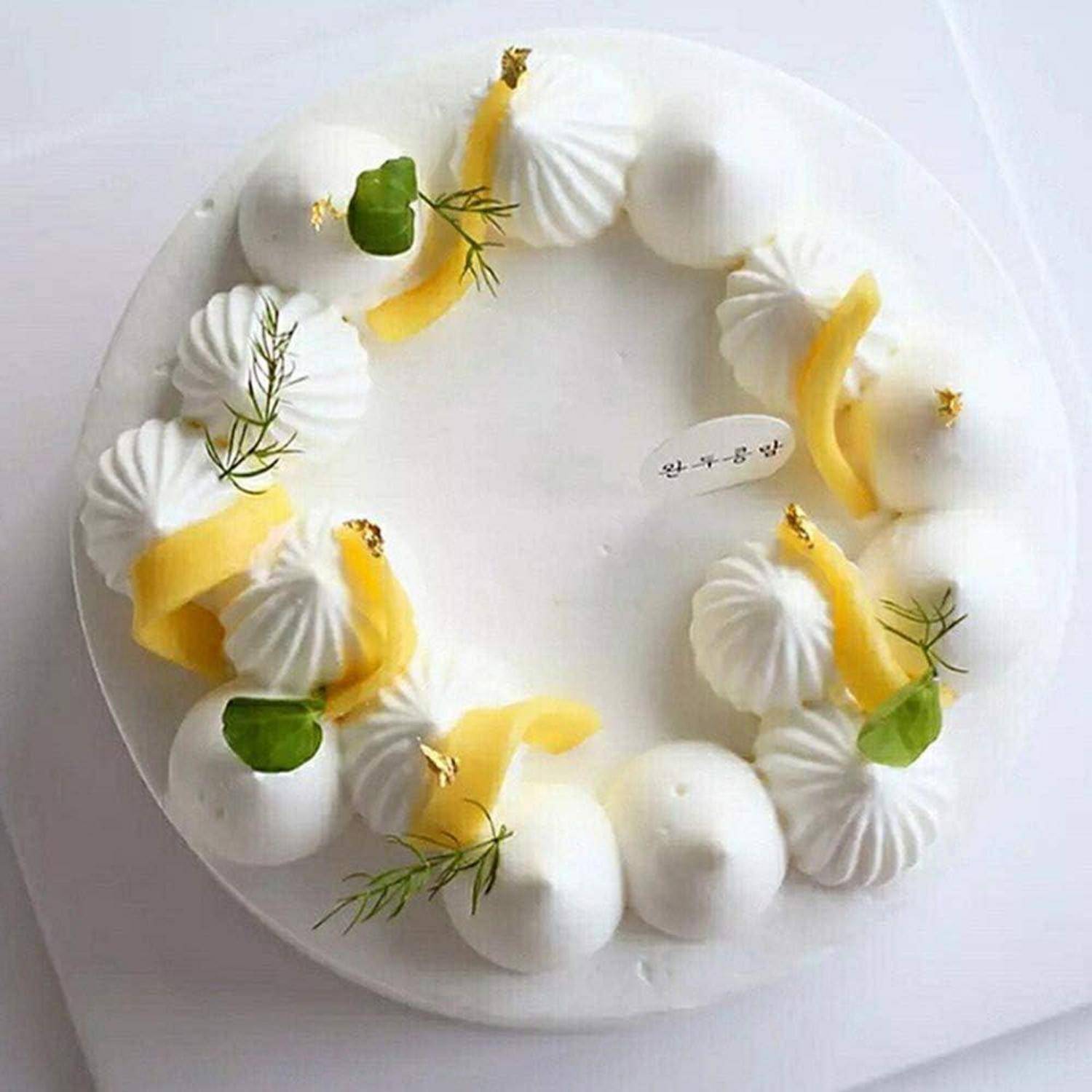 Weißer runder Kuchen, verziert mit Schlagsahnewirbeln, Mangoscheiben, Minzblättern und kleinen goldenen Akzenten auf einem weißen Teller.