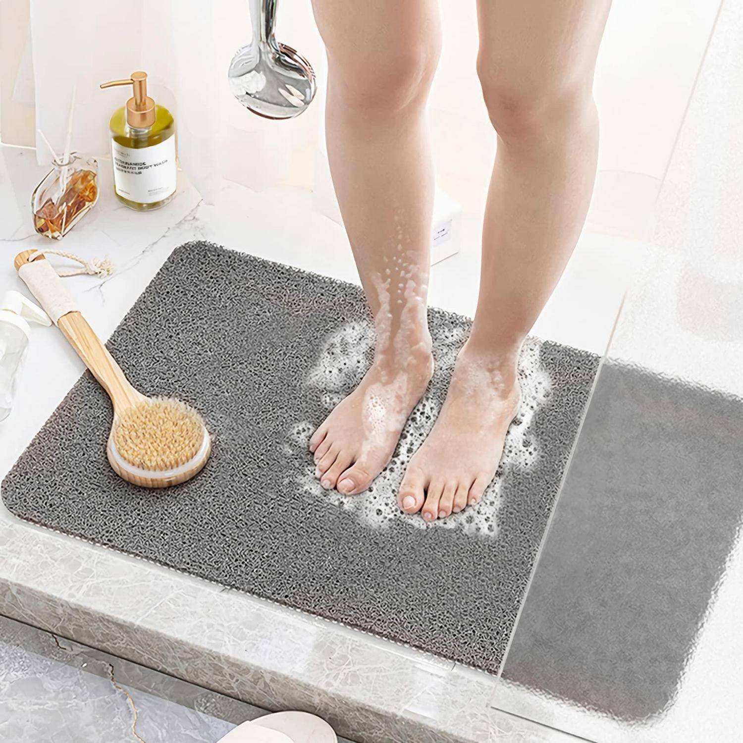 Person, die auf einer grauen Badematte duscht, mit Seifenschaum um nackte Füße, einem Duschkopf oben und einer Bürste zum Baden sowie Toilettenartikeln in der Nähe.