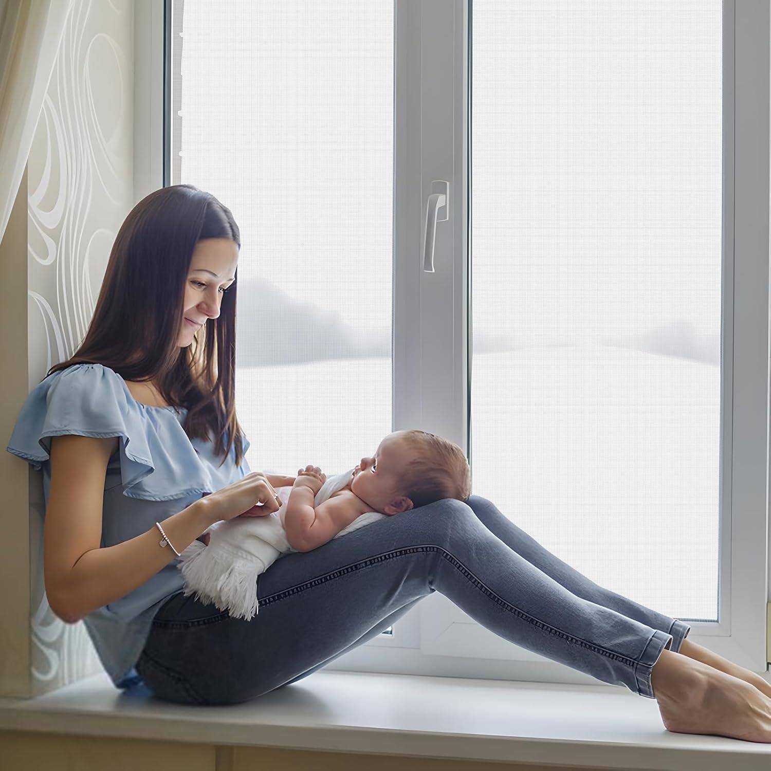 Mutter sitzt auf einer Fensterbank und hält sanft ihr Baby, das in eine weiße Decke gewickelt ist, während sie es anlächelt.