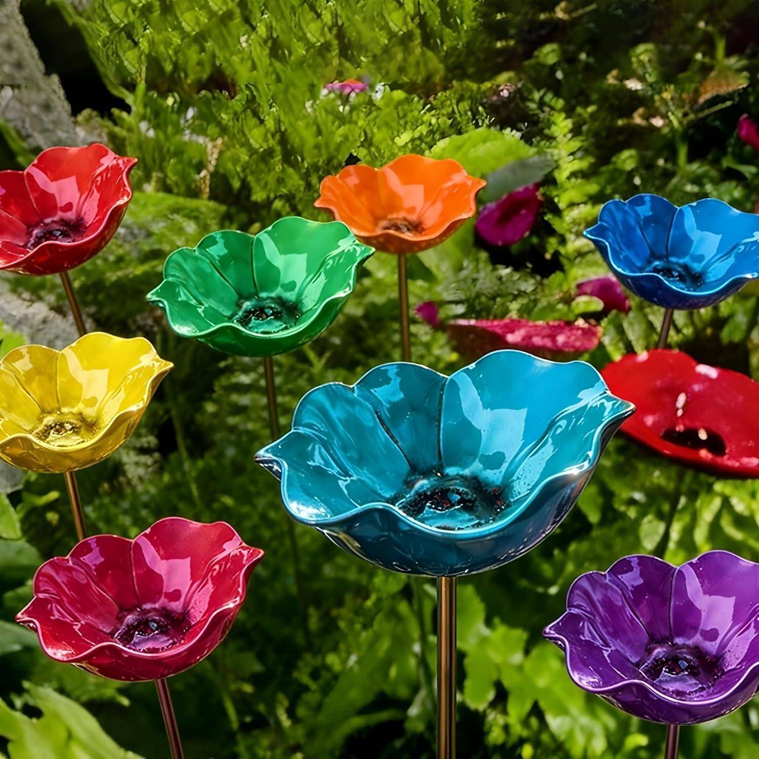 Bunte Glasblumenskulpturen auf dünnen Stielen in einem Garten, die roten, orangefarbenen, gelben, grünen, blauen, rosa und violetten Mohnblumen ähneln.