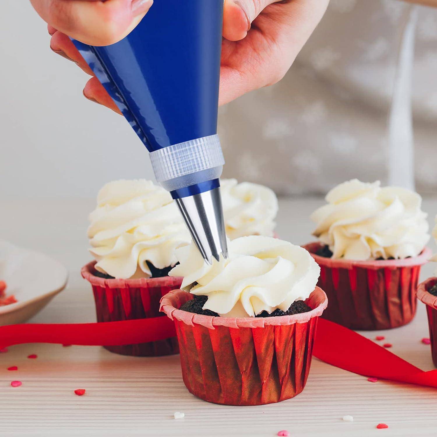Eine Hand spritzt weiße Glasur auf Schokoladen-Cupcakes in roten Förmchen, mit einem roten Band auf einem hellen Tisch.