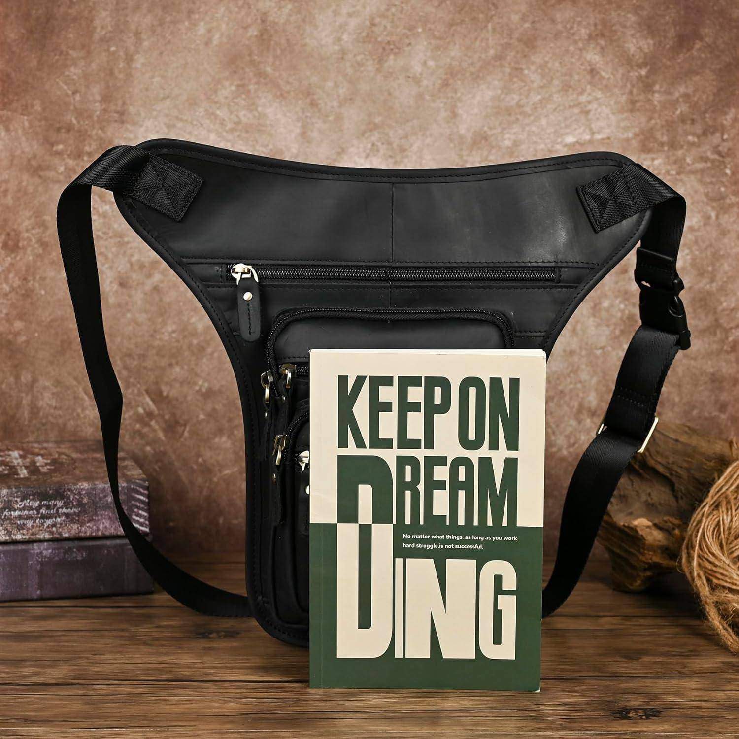 Schwarze Umhängetasche mit mehreren Reißverschlusstaschen hinter einem Buch oder einer Karte, auf der "KEEP ON DREAMING"" steht, auf einer Holzoberfläche.