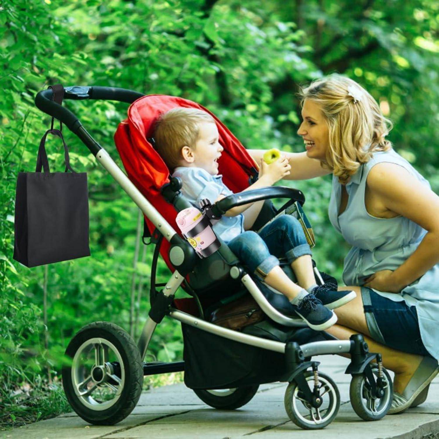 Eine Frau kniet neben einem Kinderwagen und füttert ein Kleinkind mit einem Snack auf einem bewaldeten Weg, während eine hängende Einkaufstasche am Kinderwagen befestigt ist.