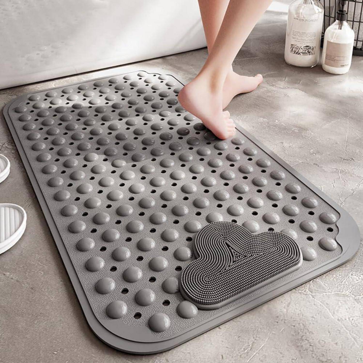 Eine Person tritt auf eine graue strukturierte Badmatte mit erhabenen Noppen und einem kleinen fußförmigen Schrubbpad in einem Badezimmer.