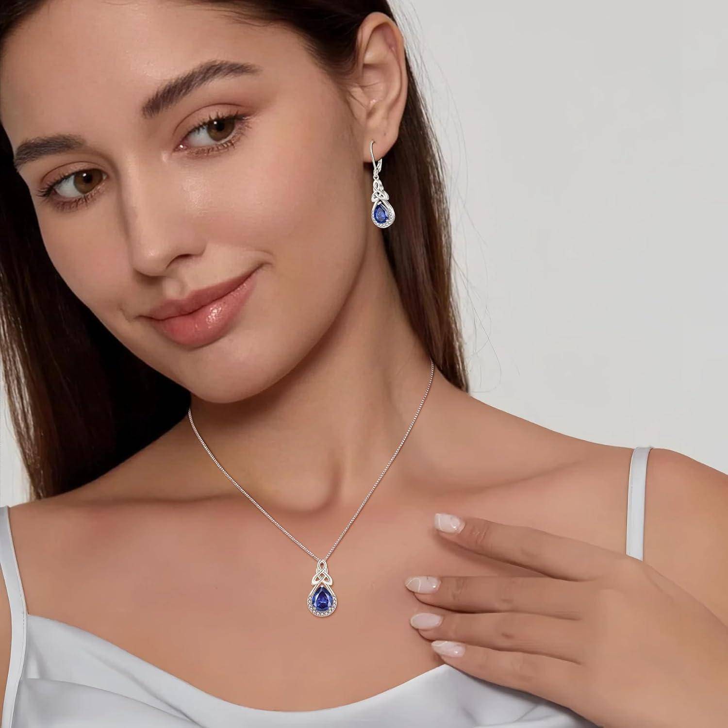 Porträt einer Frau, die eine zusammengehörige Halskette mit blauem Edelsteinanhänger und Tropfenohrringe vor einem einfachen Hintergrund modelt.