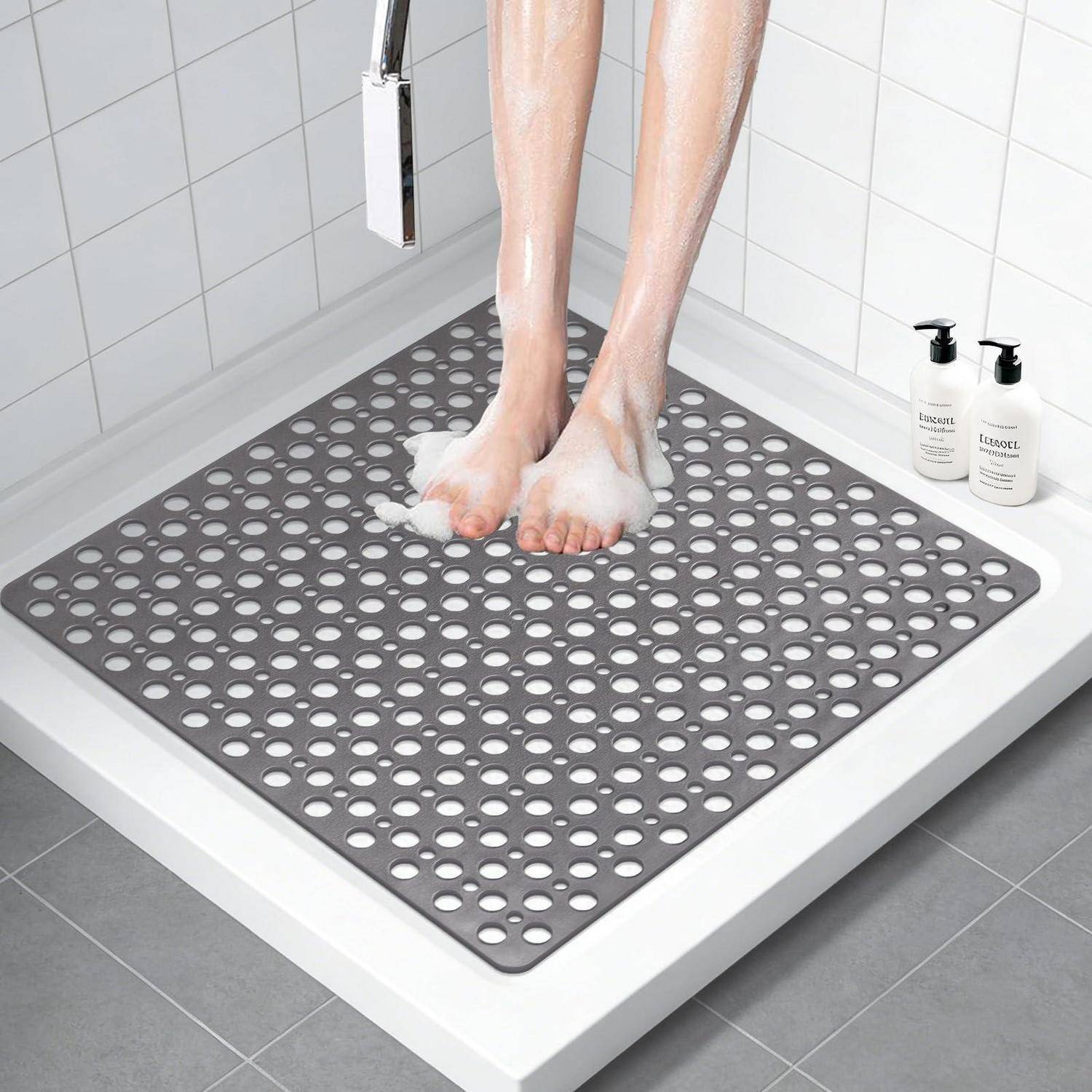 Person, die auf einer grauen perforierten Badmatte in einer weißen gefliesten Dusche duscht, mit zwei Pumpflaschen auf dem Regal.