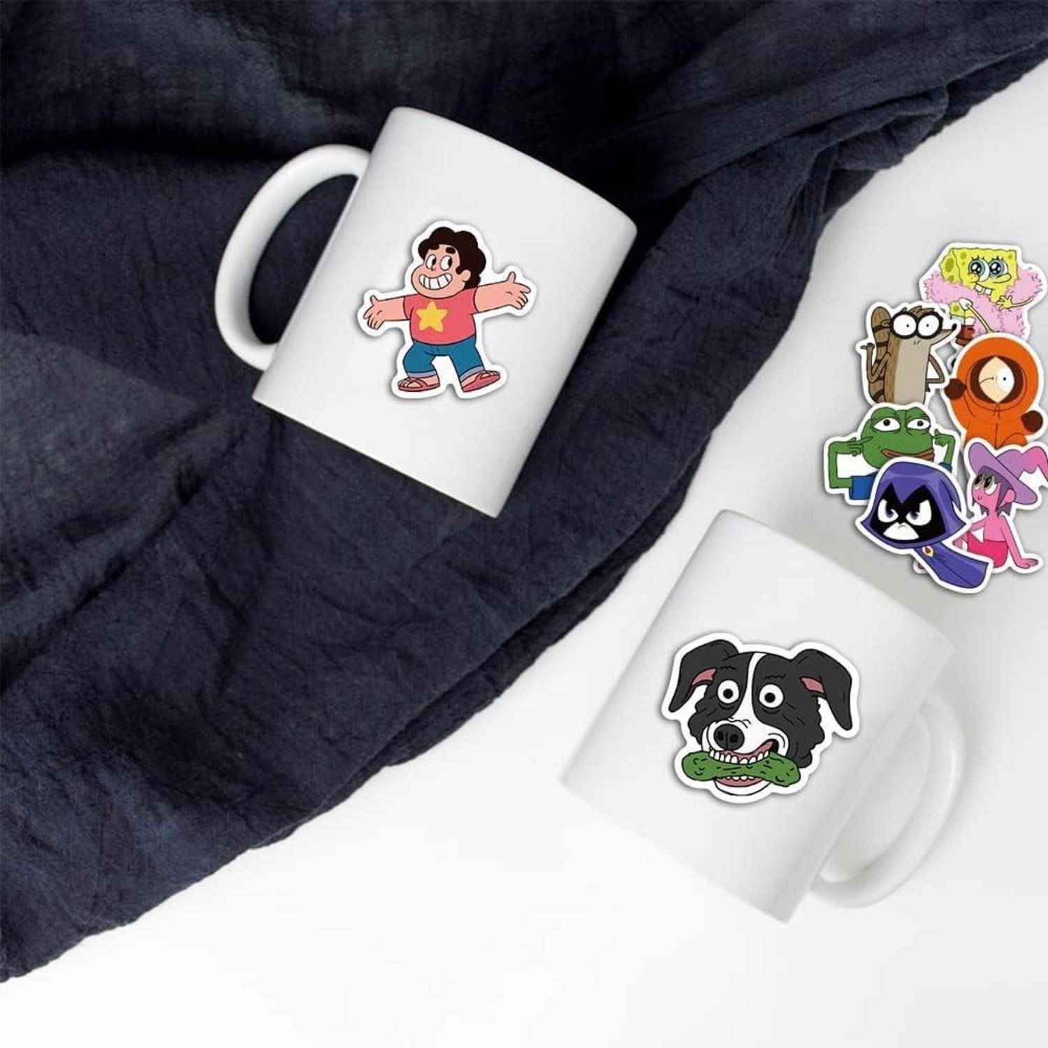 Zwei weiße Tassen mit Aufklebern von Cartoon-Charakteren auf einem dunklen Stoff-Hintergrund, mit zusätzlichen Charakteraufklebern, die auf der rechten Seite angeordnet sind.