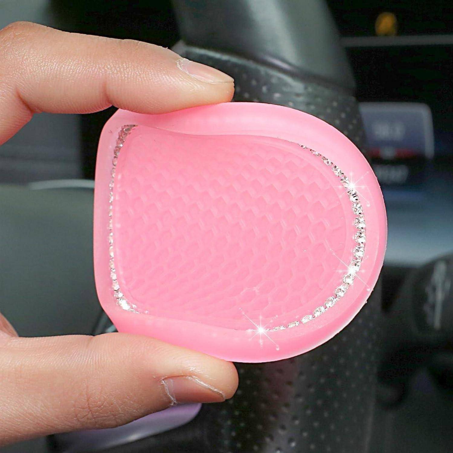 Hand, die einen rosa, herzförmigen strukturierten Autolufterfriscer mit glitzerndem Rand in einem Fahrzeug hält.