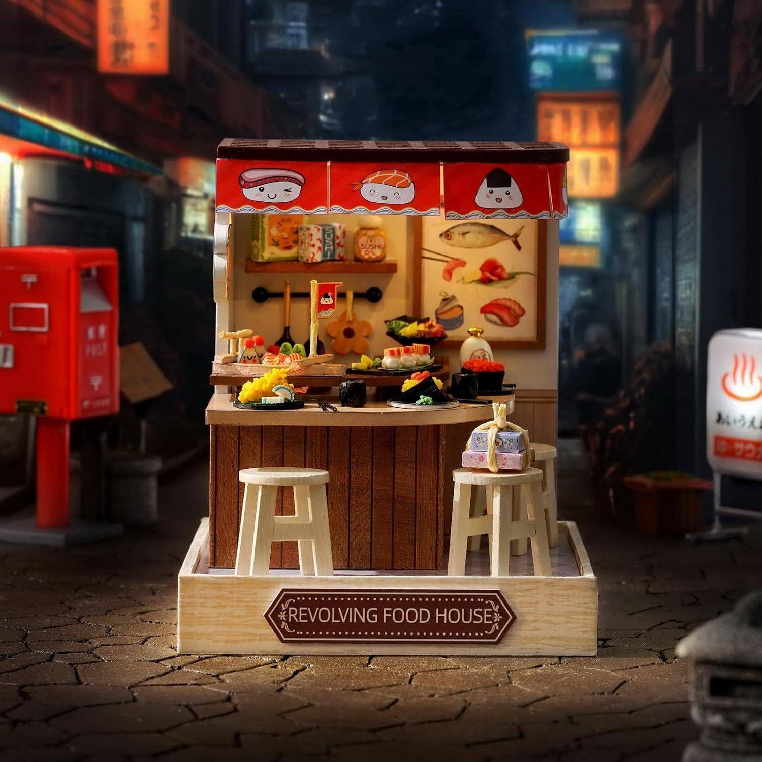 Miniatur-"Revolving Food House"" Sushi-Verkaufsstand mit Hockern, Lebensmittelschalen und rotem Markisen in einer dunklen Straßenszene.