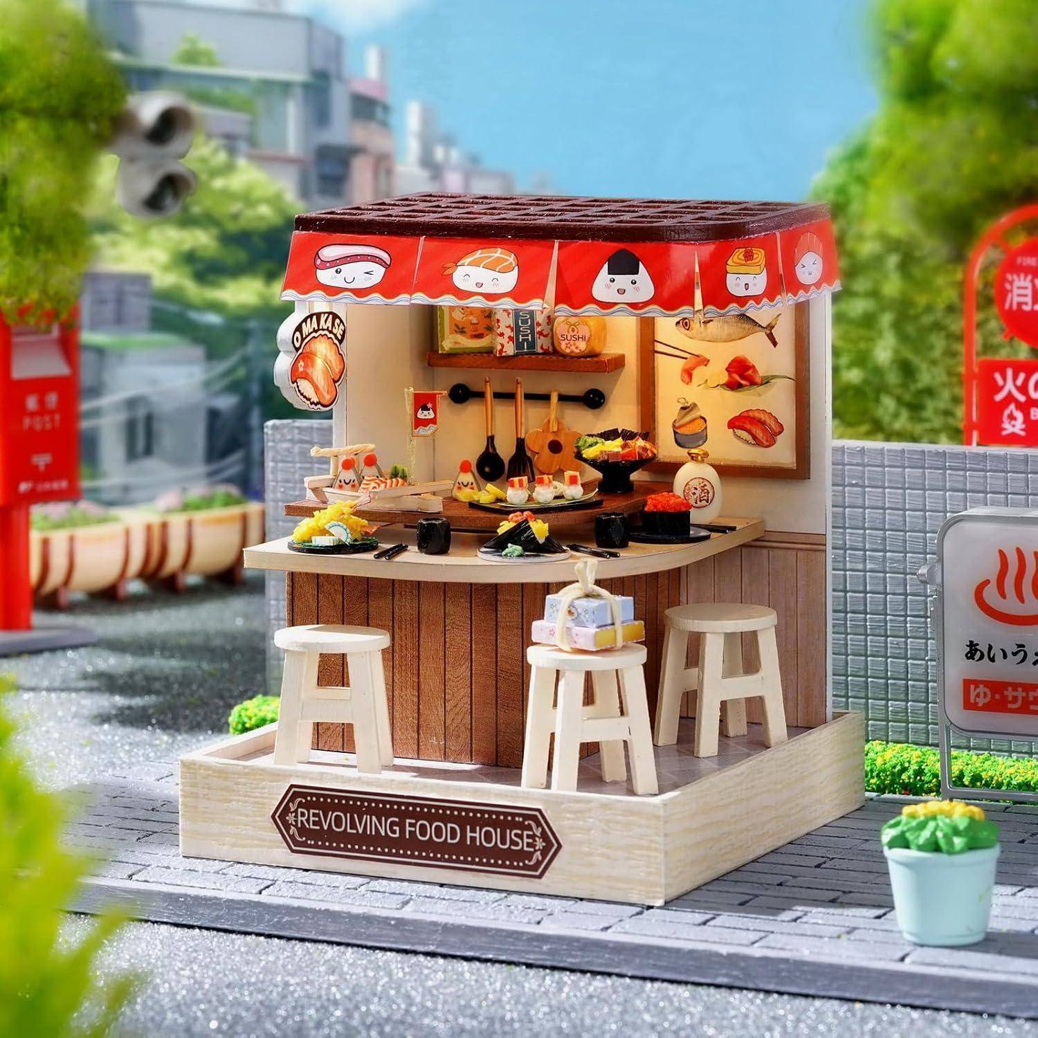 Miniatur-Essensstand mit der Bezeichnung „REVOLVING FOOD HOUSE" mit Sushi- und japanischen Snack-Auslagen, Hockern und einer roten Markise auf einer Stadtstraße.