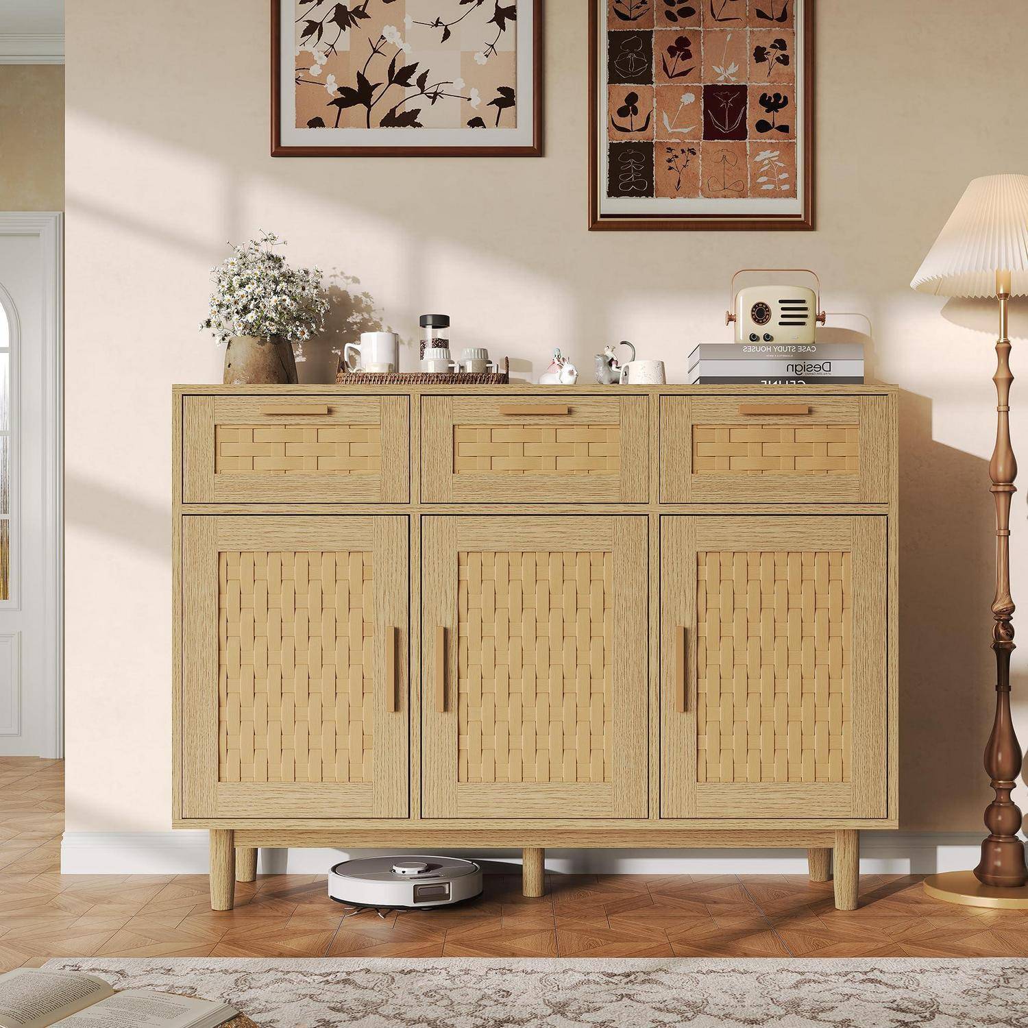Ein helles Holzkommode mit gewebten Schranktüren steht gegen eine beige Wand, dekoriert mit Dekoration und gerahmter Kunst, mit einem Roboterstaubsauger darunter.