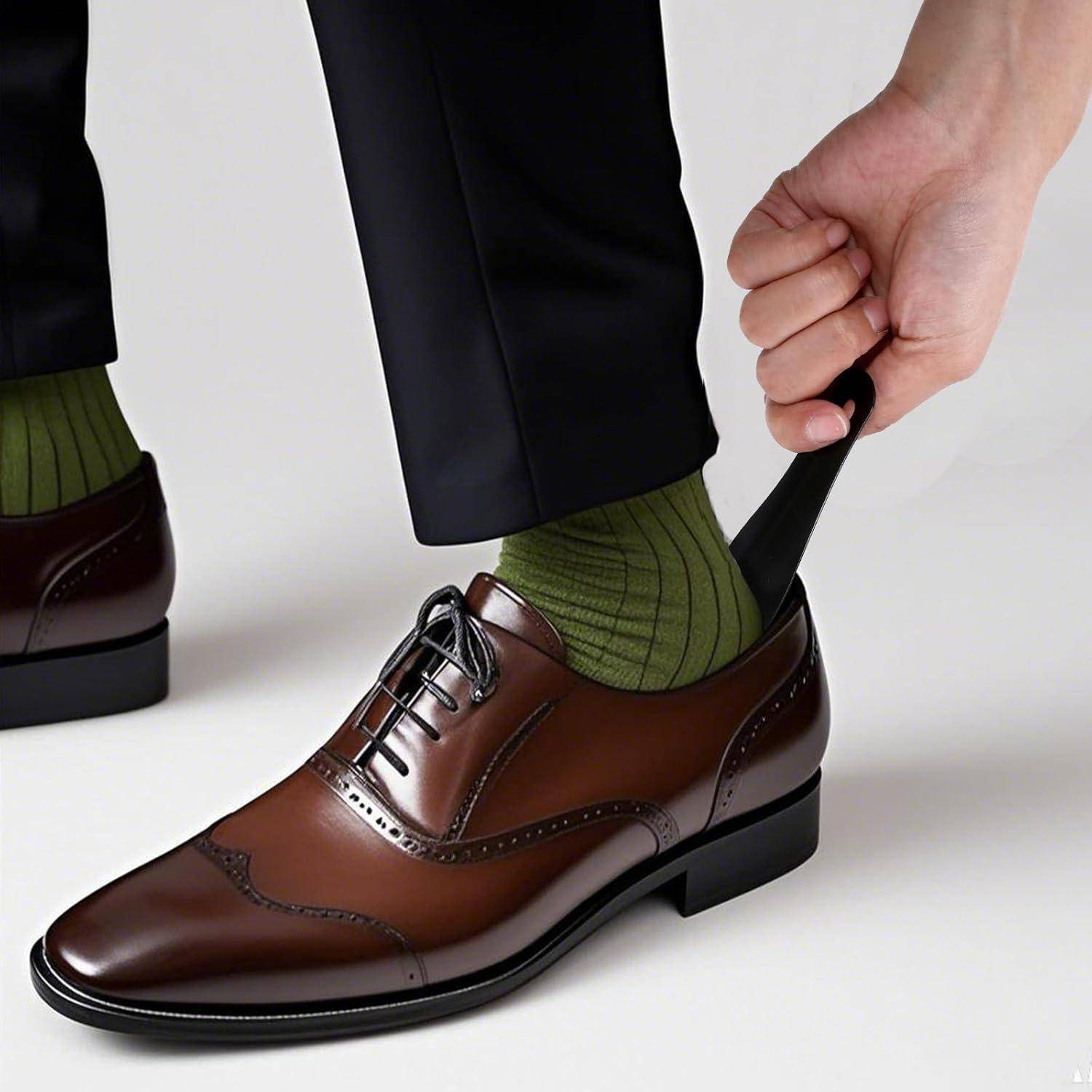 Eine Hand passt eine grüne Socke an, während man einen braunen Lederschuh anzieht, dazu schwarze Hosen auf weißem Hintergrund.