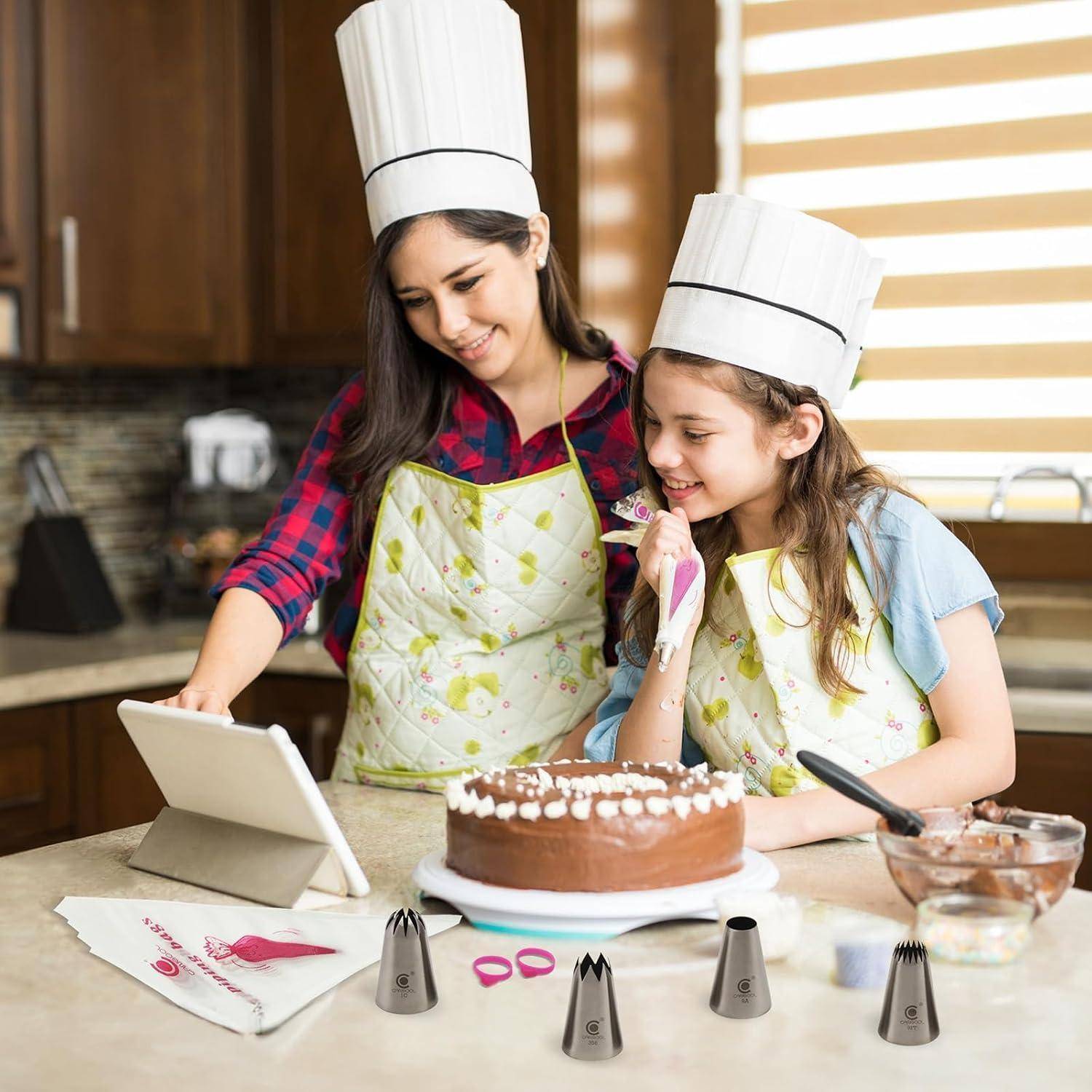 Frau und Mädchen mit Kochmützen dekorieren einen Schokoladenkuchen in einer Küche, schauen auf ein Tablet, während Zuckerguss und Werkzeuge auf der Arbeitsfläche liegen.