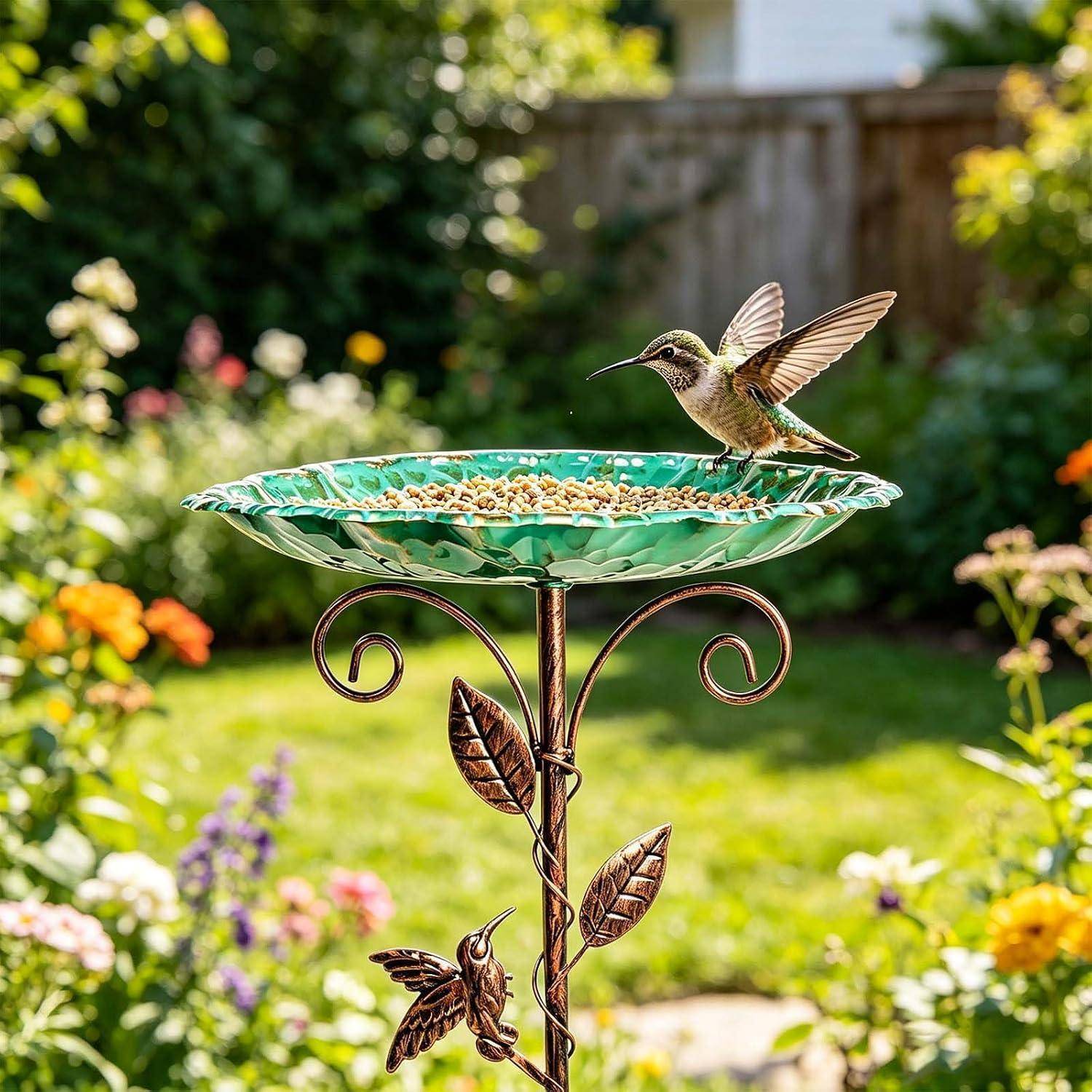 Kolibri sitzt auf einem dekorativen Vogelfutterhaus in einem Garten, mit erhobenen Flügeln, während er sich von der mit Samen gefüllten Schale ernährt.