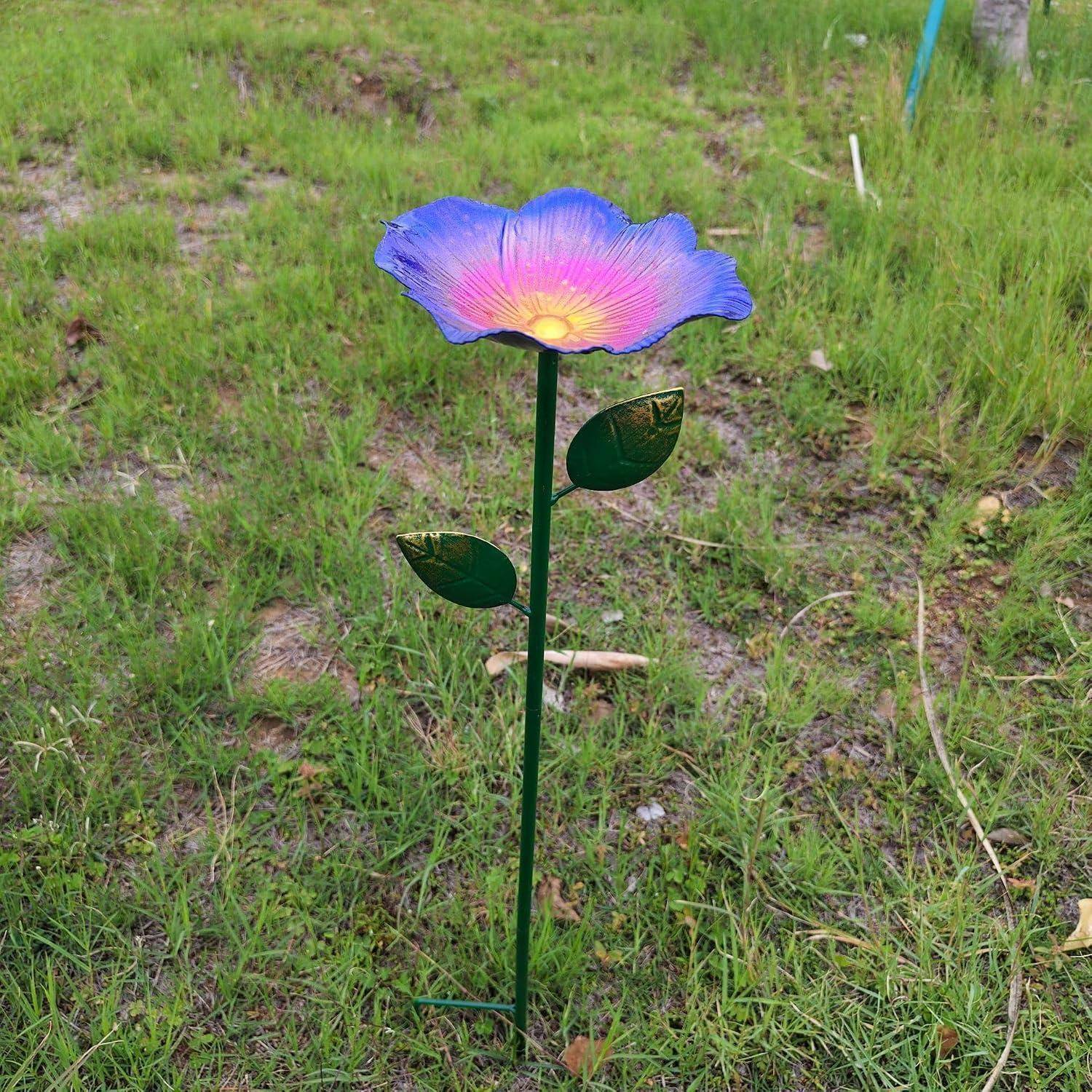 Ein dekorativer Metallblumenstab mit einer purpur-blauen Blüte und zwei grünen Blättern steht in einem grasbewachsenen Garten.