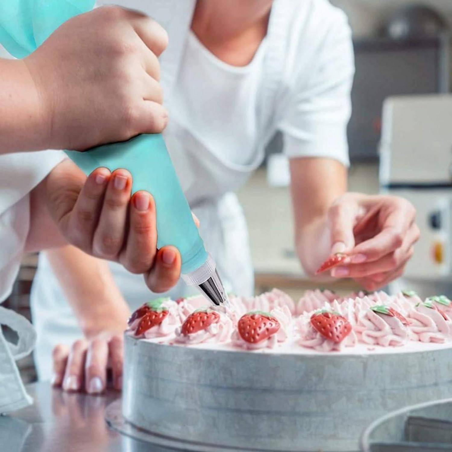 Eine Person, die rosa Glasur auf einen Kuchen mit Erdbeeren spritzt, während eine andere Hand eine Erdbeere platziert, in einer Küchen- oder Bäckerei-Umgebung.